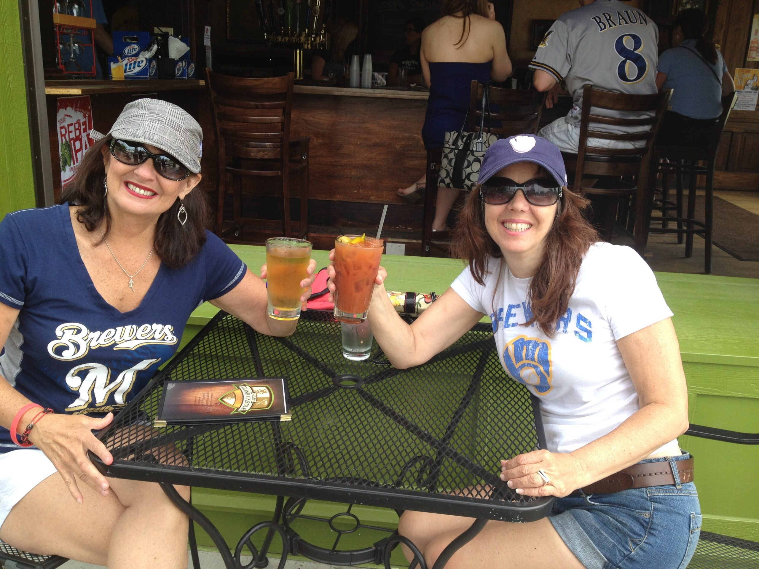 Two smiling women toast iced drinks at an outdoor cafe, wearing sunglasses; one in a Brewers shirt, the other in a white tee and baseball cap at a green table.