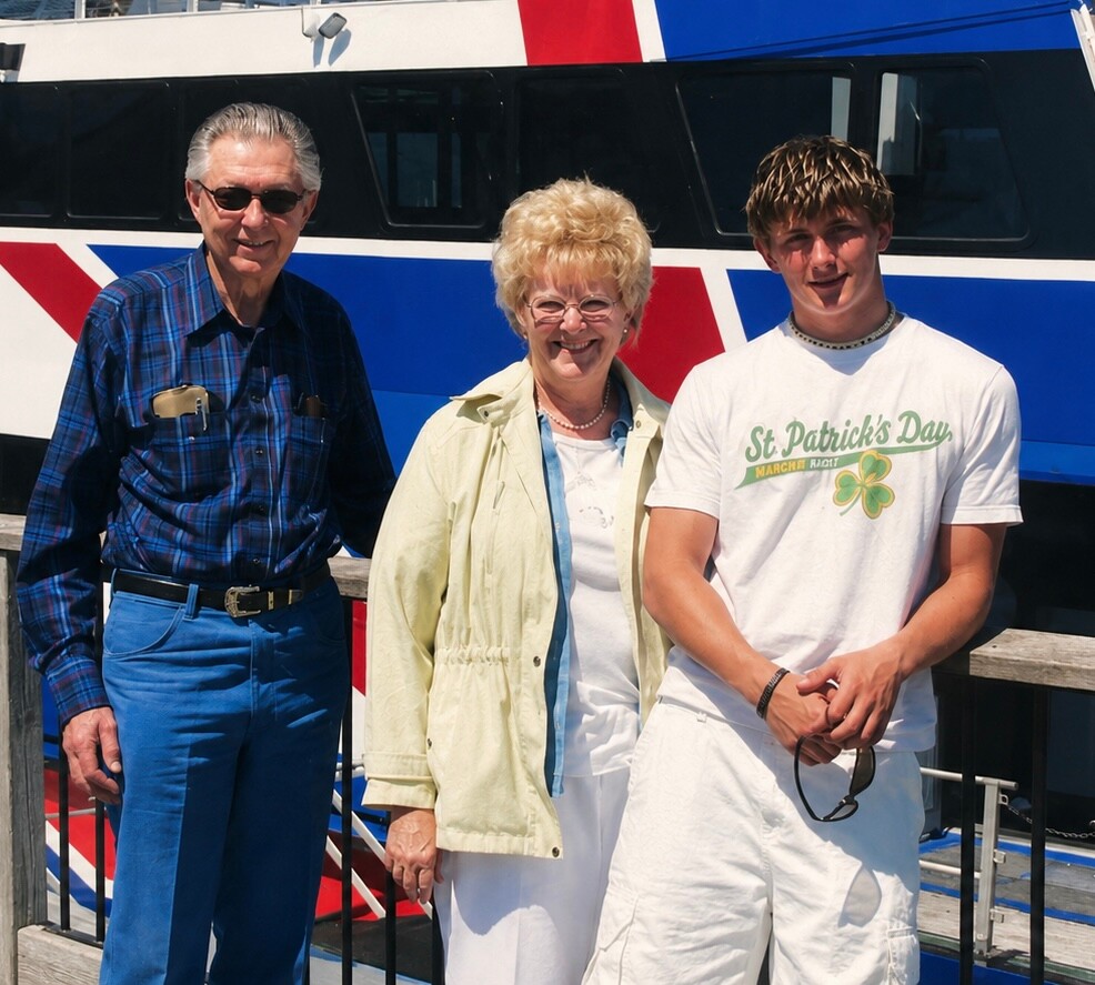Three people posing in front of a colorful train: an older man, a smiling woman, and a young man in a St. Patrick’s tee.