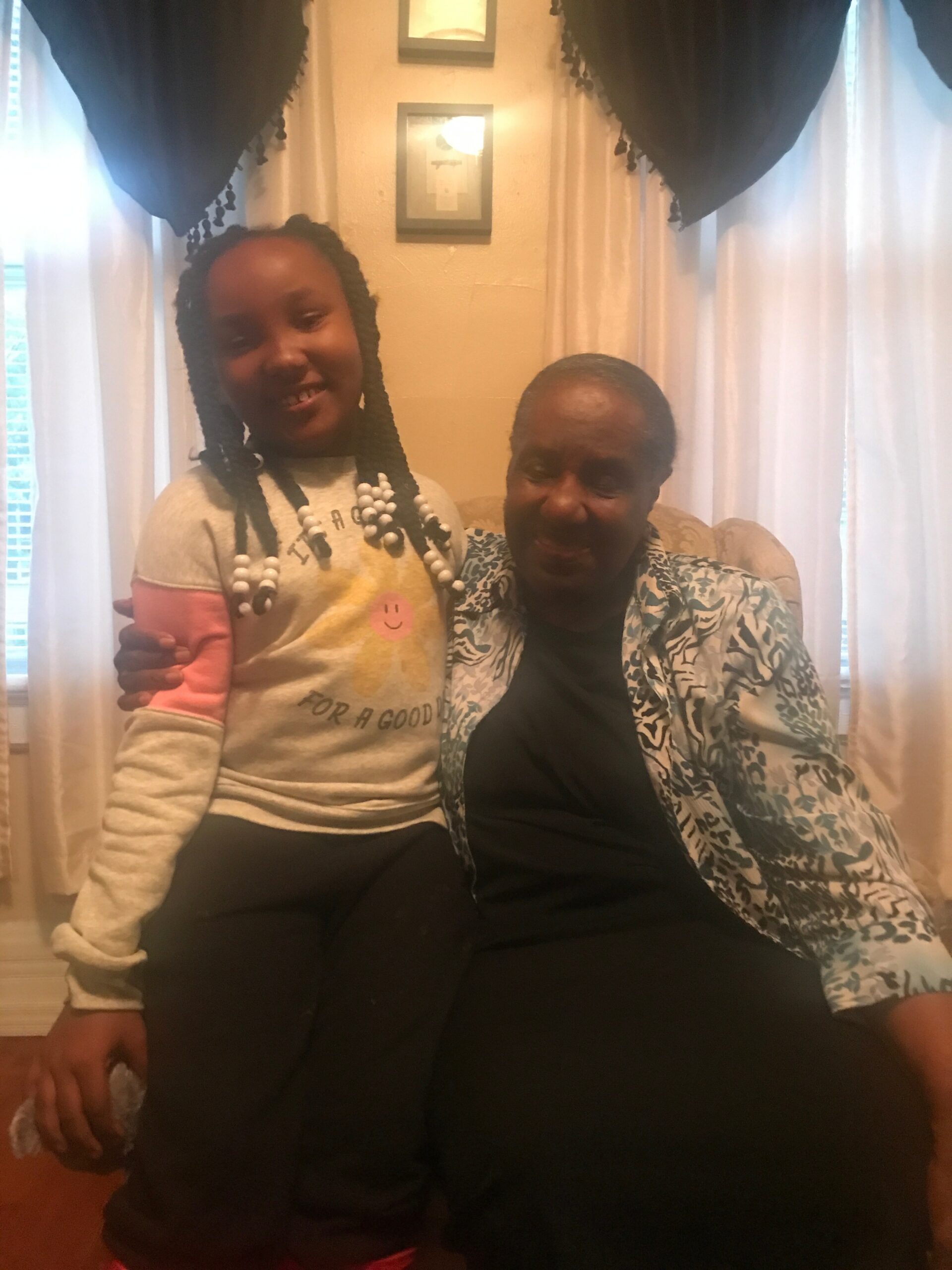Smiling girl with braids and beads sits beside an elderly woman in a patterned jacket on a couch in a living room.