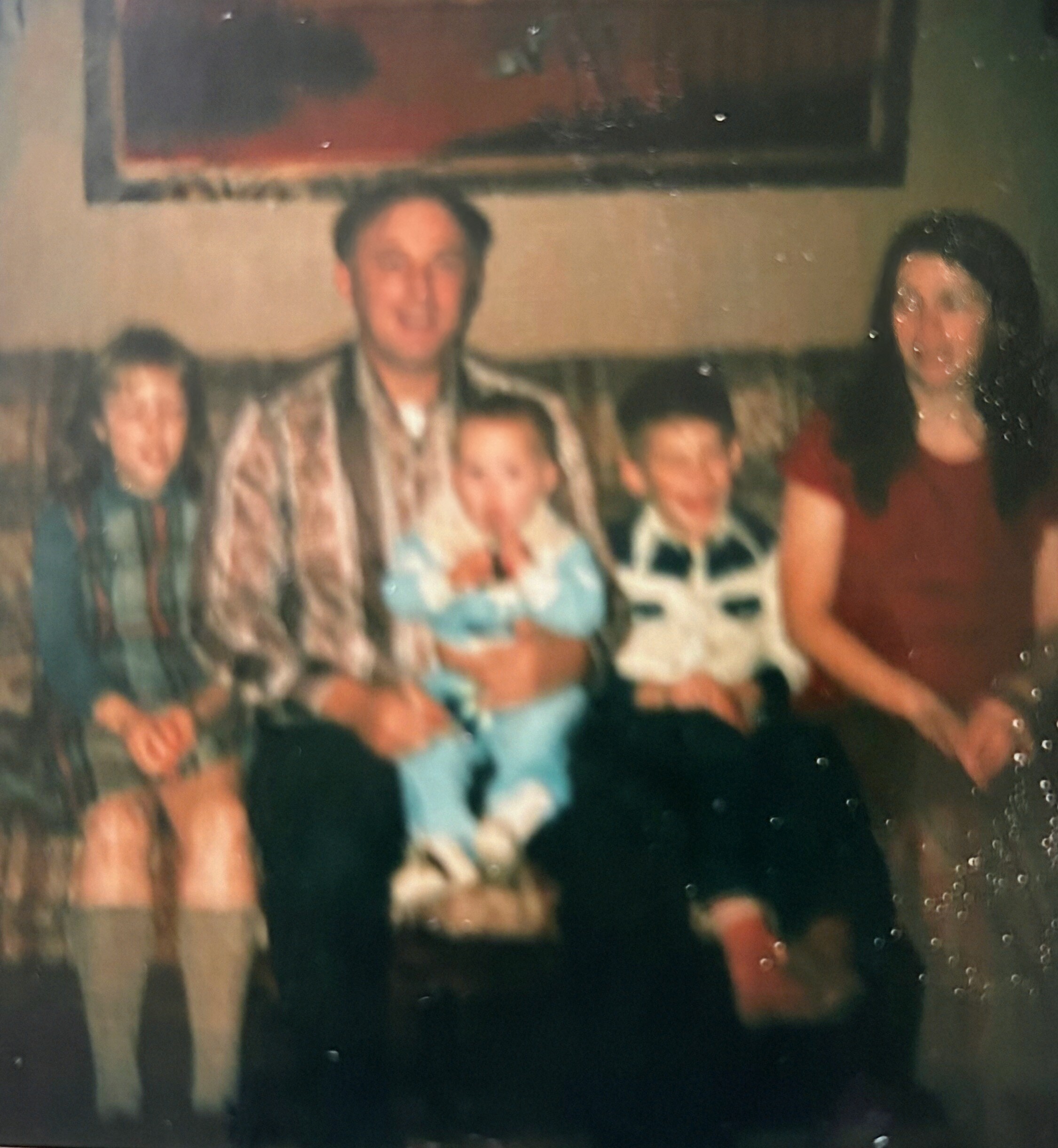 Blurry indoor group photo of adults and children posing together at a social gathering, with a framed picture on the wall in the background.