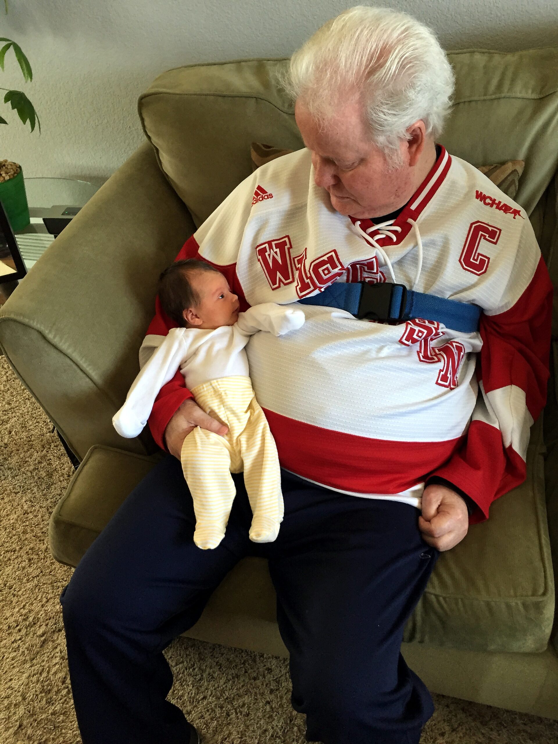 Older man in a red-and-white hockey jersey cuddling a newborn baby wrapped in a yellow blanket on a sofa.