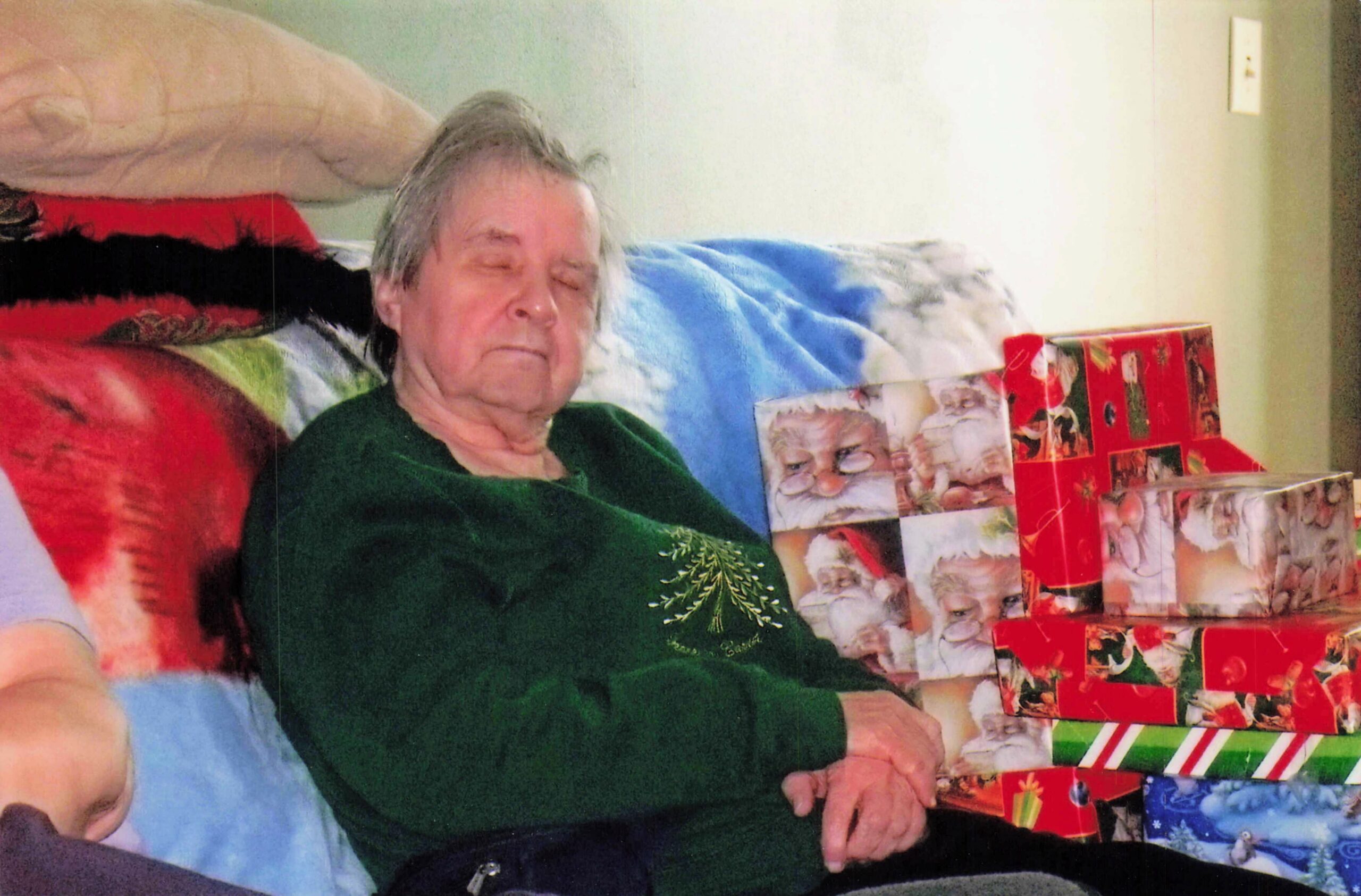 Older woman in a green sweater sits with eyes closed among wrapped Christmas presents on a couch.