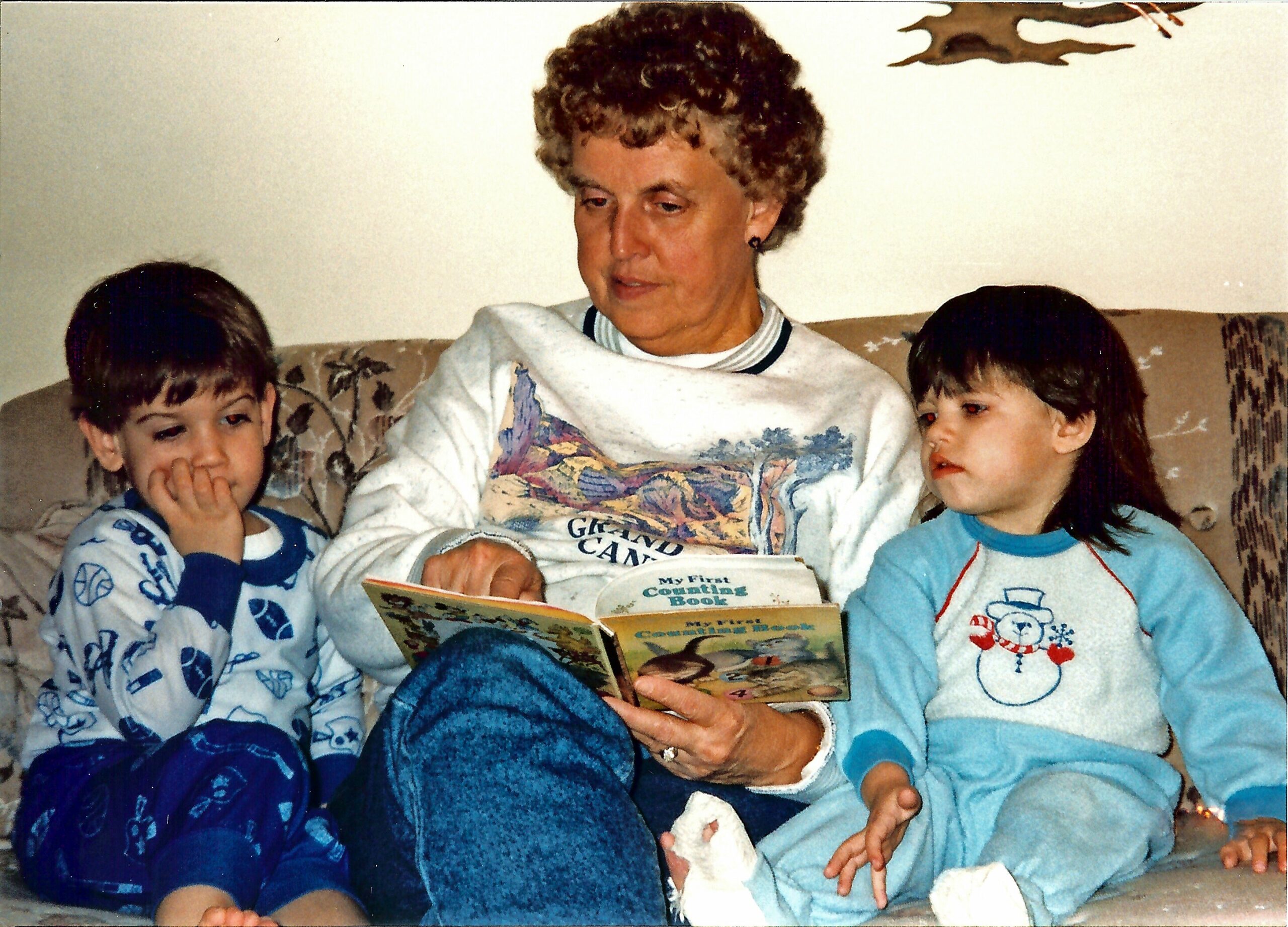 Older woman with short curly hair reading aloud from a children's book to two young children sitting on a couch in pajamas; book title visible.”,