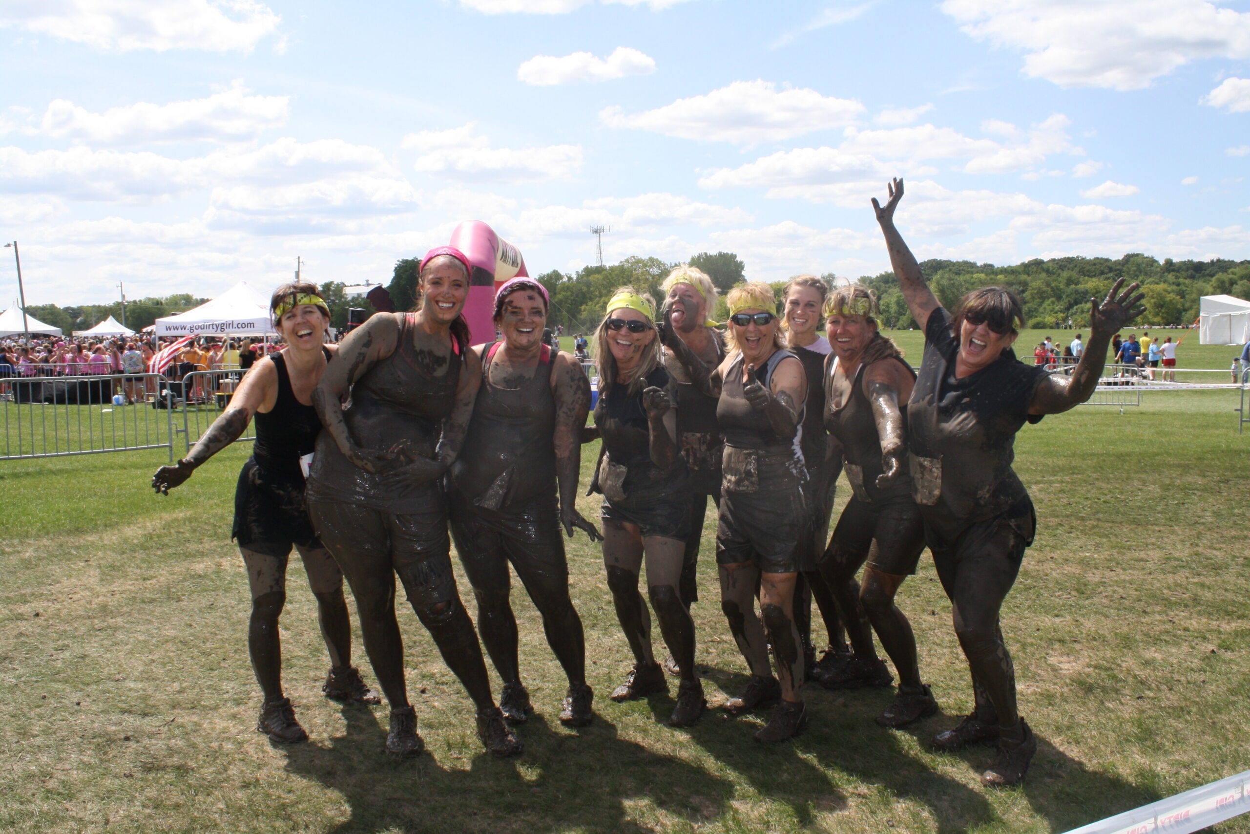 Group of women covered in mud posing and smiling at an outdoor obstacle event in a park.