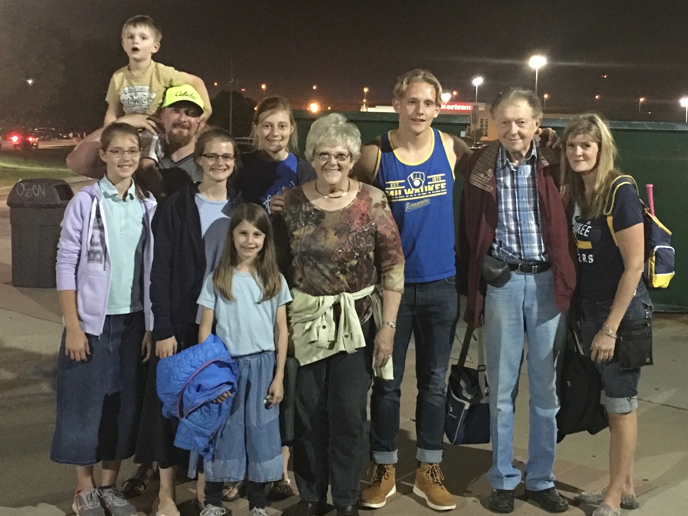 Group of people posing for a photo at night outside, including a child on someone's shoulders and several adults smiling. Behind them are illuminated streetlights and a dark sky.
