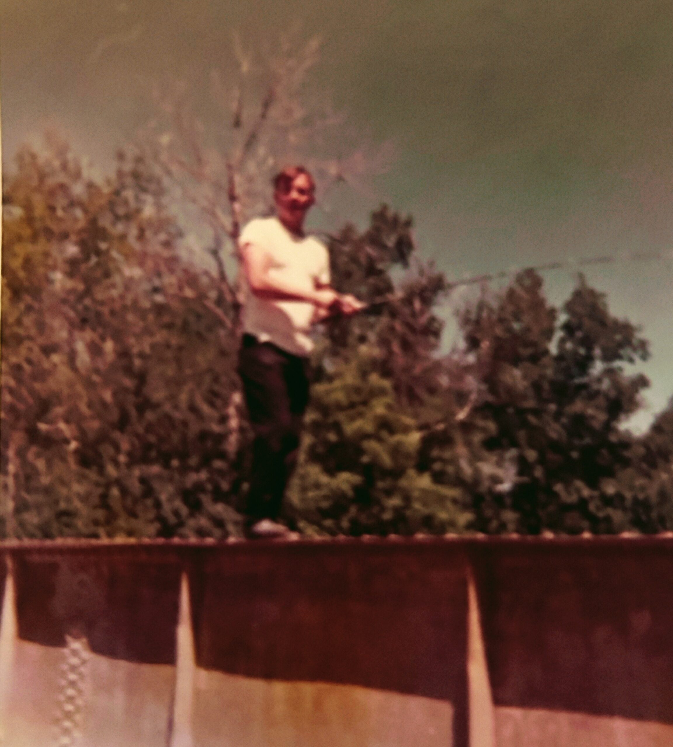 Man wearing a light shirt stands outdoors, holding a fishing rod among bushes and trees in a blurry photo.
