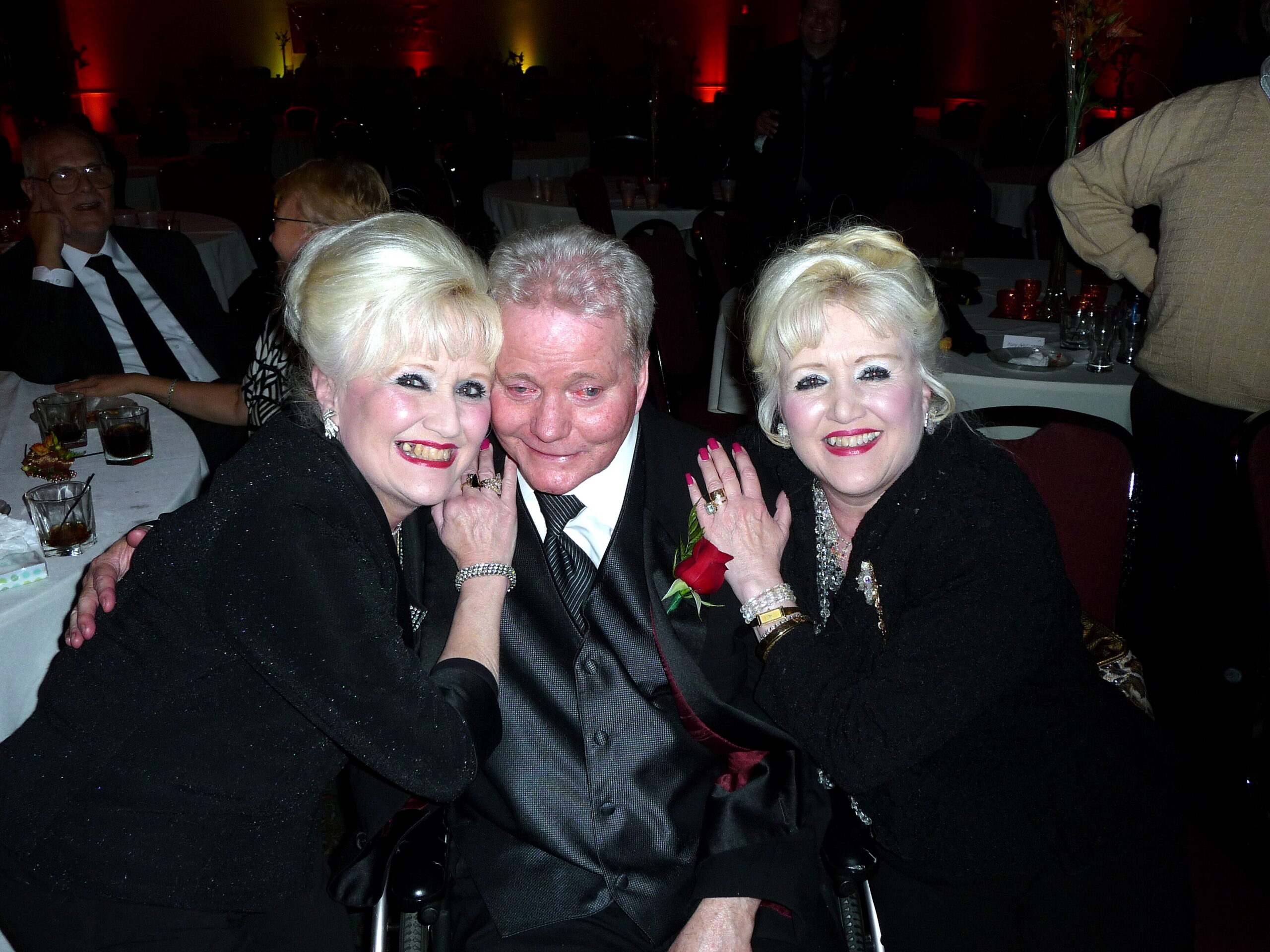 Three guests in formal attire smiling for a photo at a banquet, drinks on tables behind them.