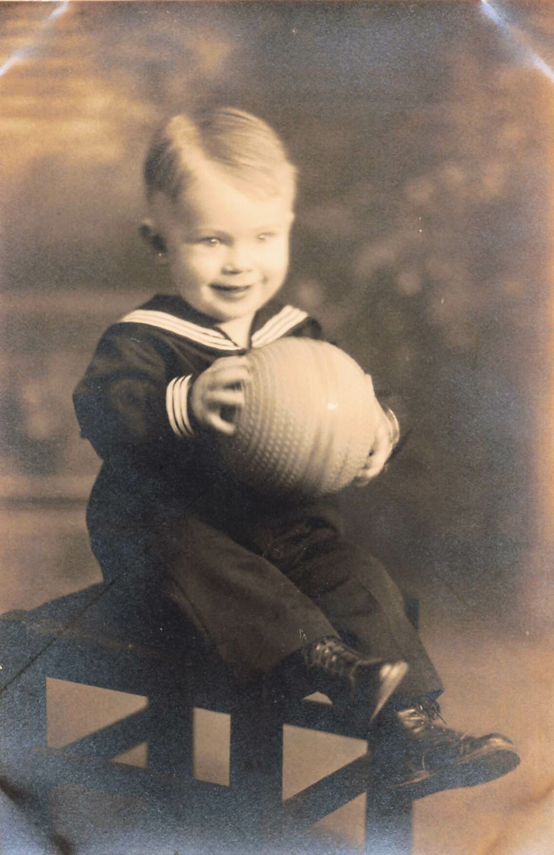 Young child in a sailor outfit sits on a wooden fence, smiling while offering a striped ball to the camera.