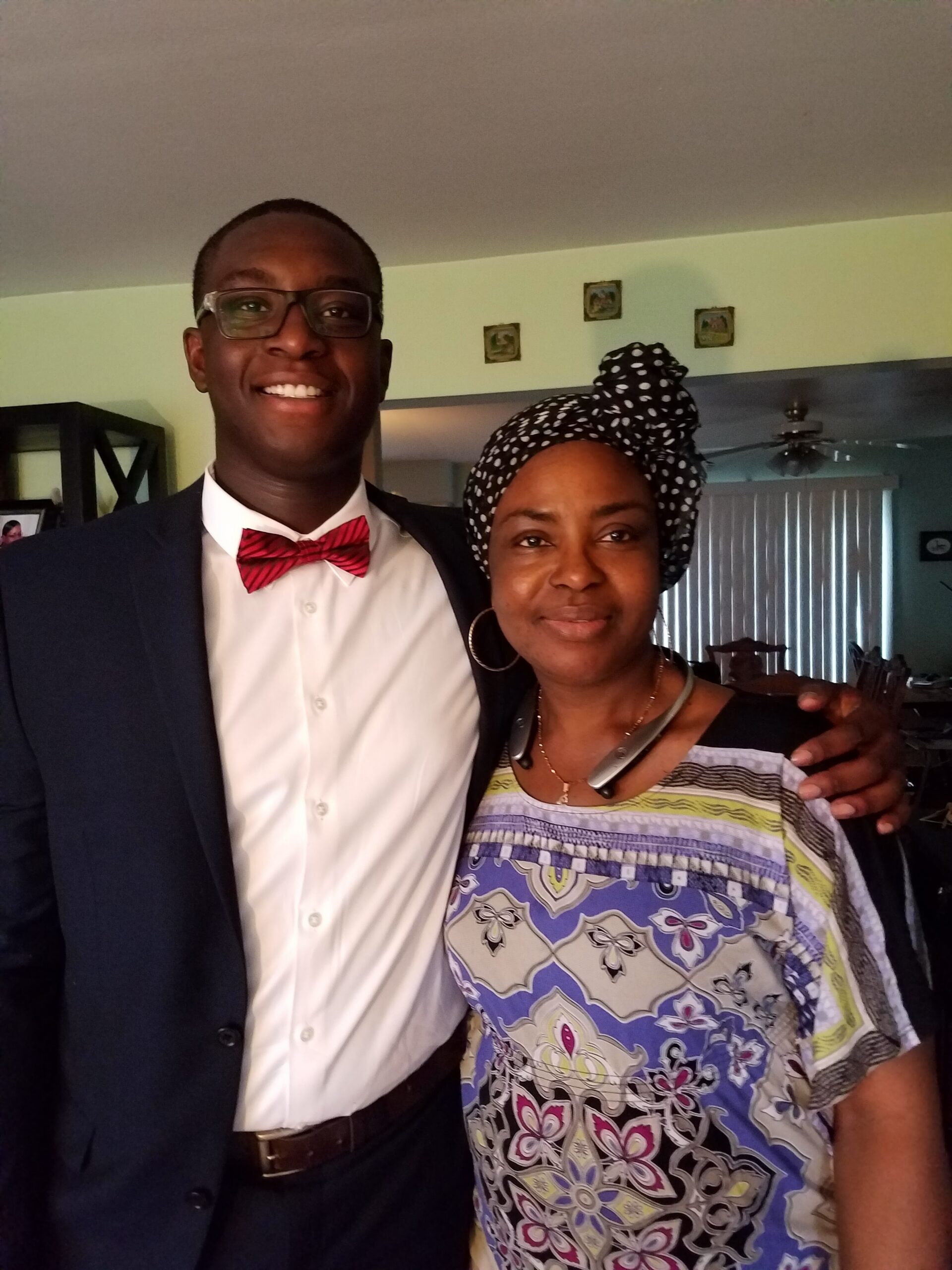Two adults posing together indoors: man in suit with red bow tie and glasses, woman in colorful patterned top with a black polka-dot headwrap, smiling.
