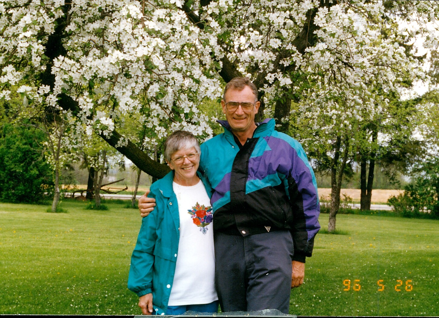 Smiling elderly couple stands with arms around each other under a blossoming white-flowered tree in a park.