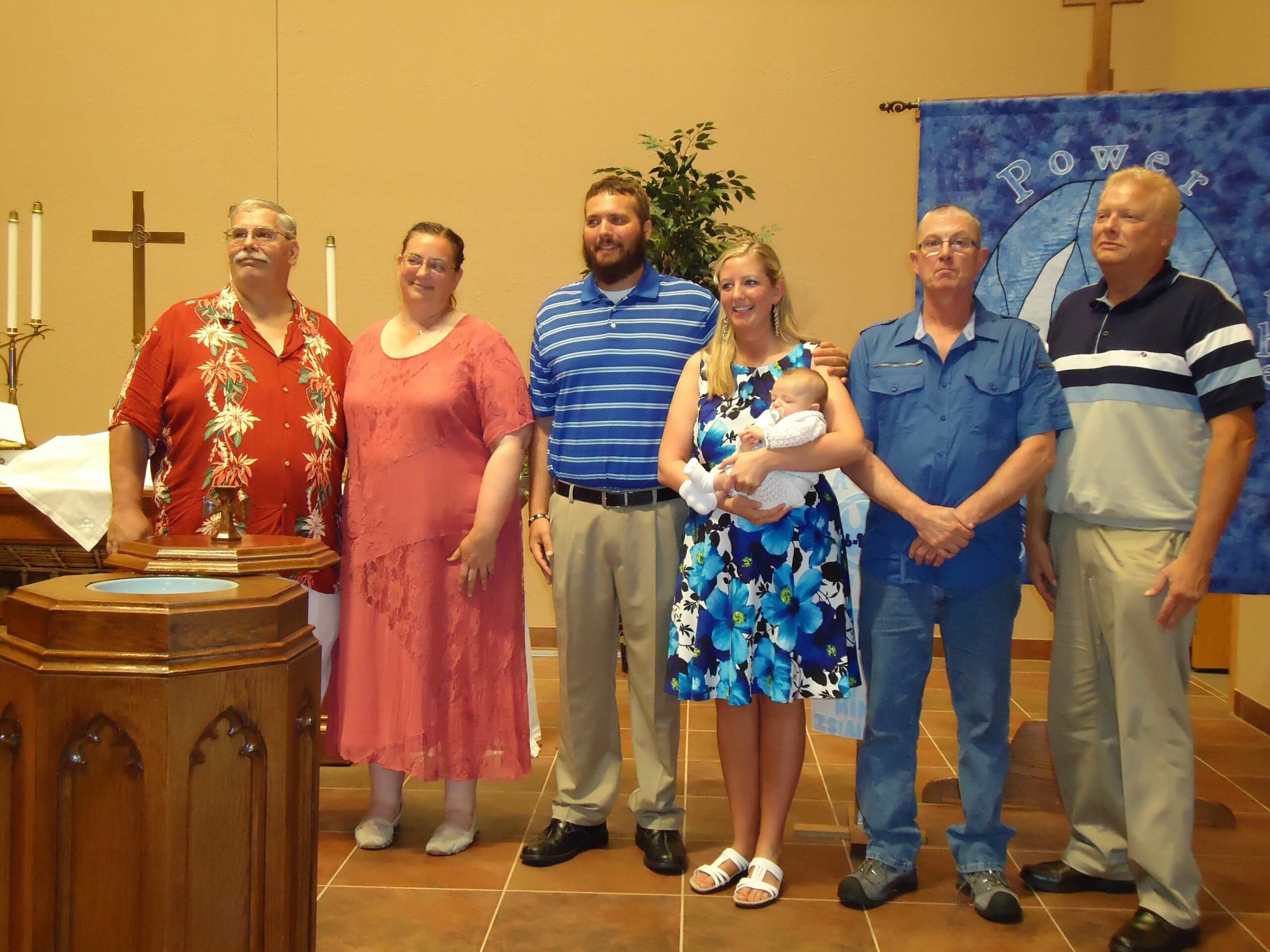 Group of adults and a baby posing for a photo inside a church, standing near a baptismal font and a cross with candles in the background.