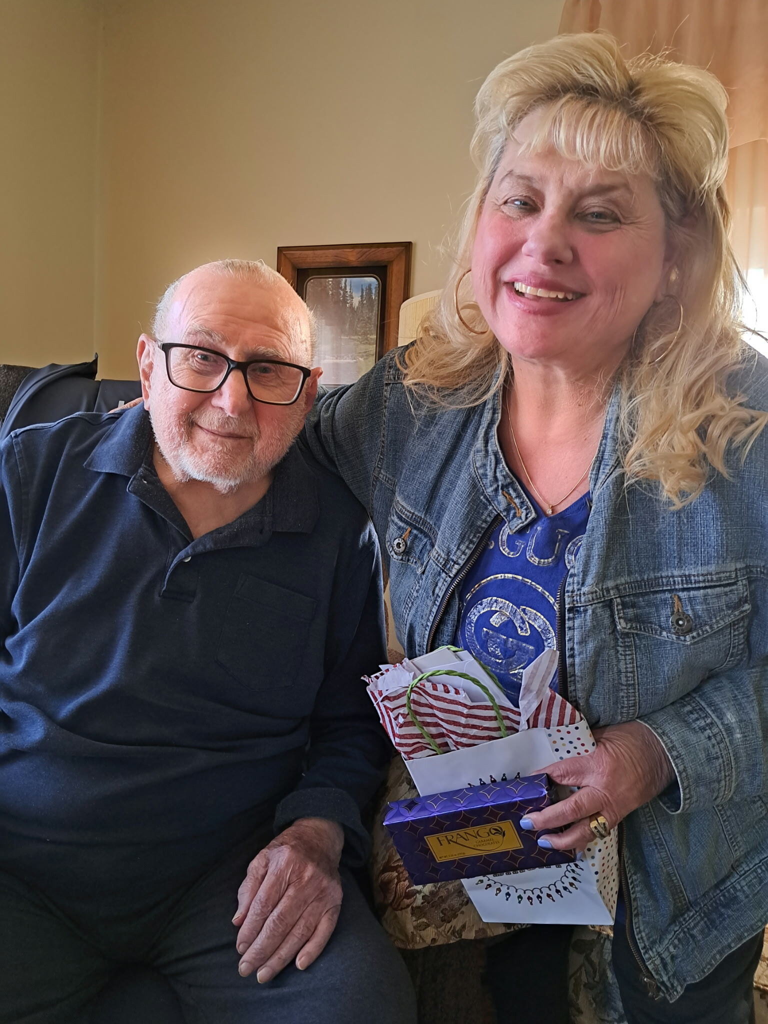 Smiling elderly man in a dark polo with glasses sits beside a smiling woman holding wrapped gifts in a cozy room.