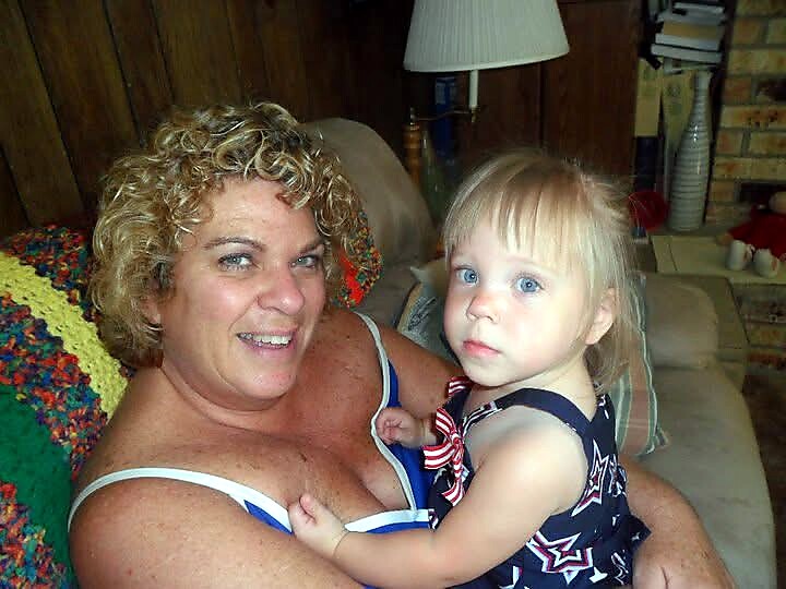 Older woman with curly hair sits on a couch, smiling, holding a young blonde girl in a navy star-pattern dress.