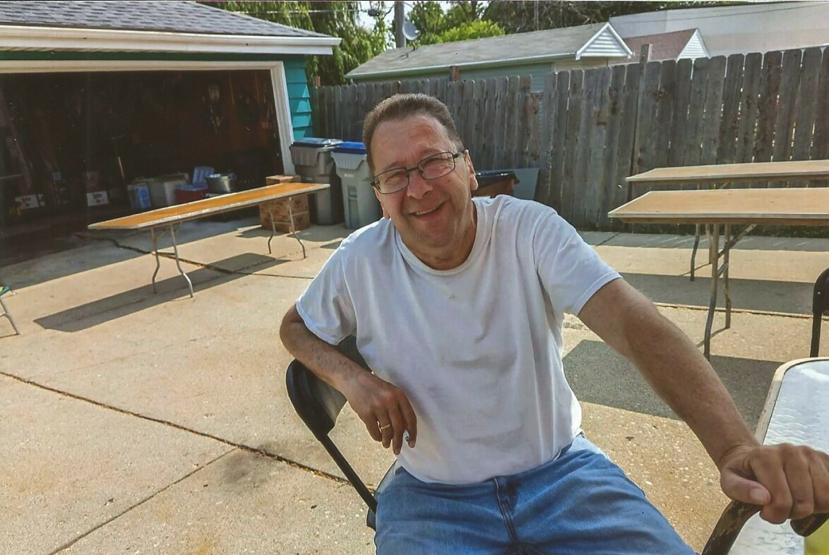 Smiling man in a white T-shirt and blue jeans sits on a chair in a sunny backyard with picnic tables and a wooden fence in the background.