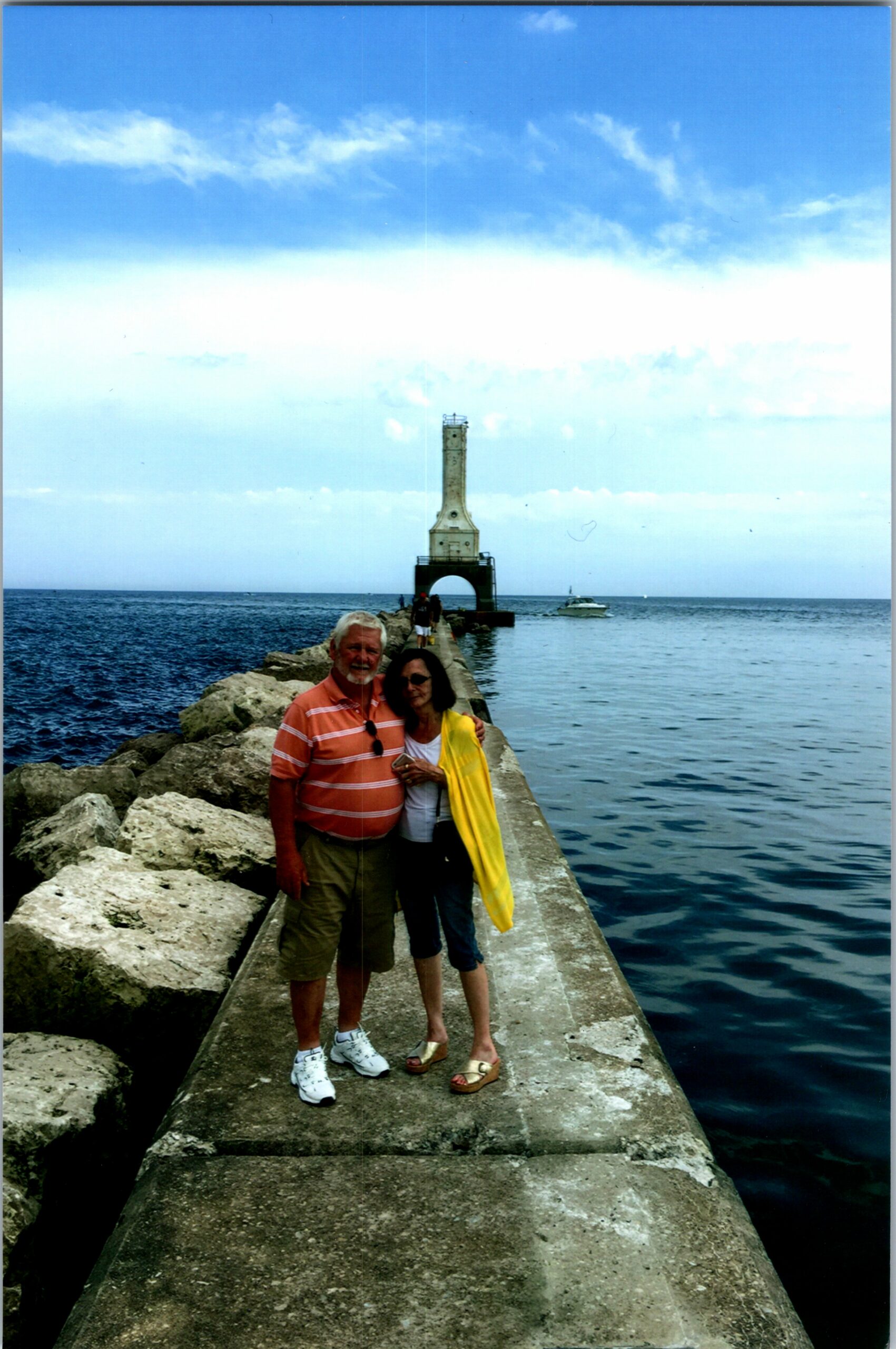 Couple posing on a concrete jetty extending into the sea, with a tall lighthouse at the end and a boat in the distance.