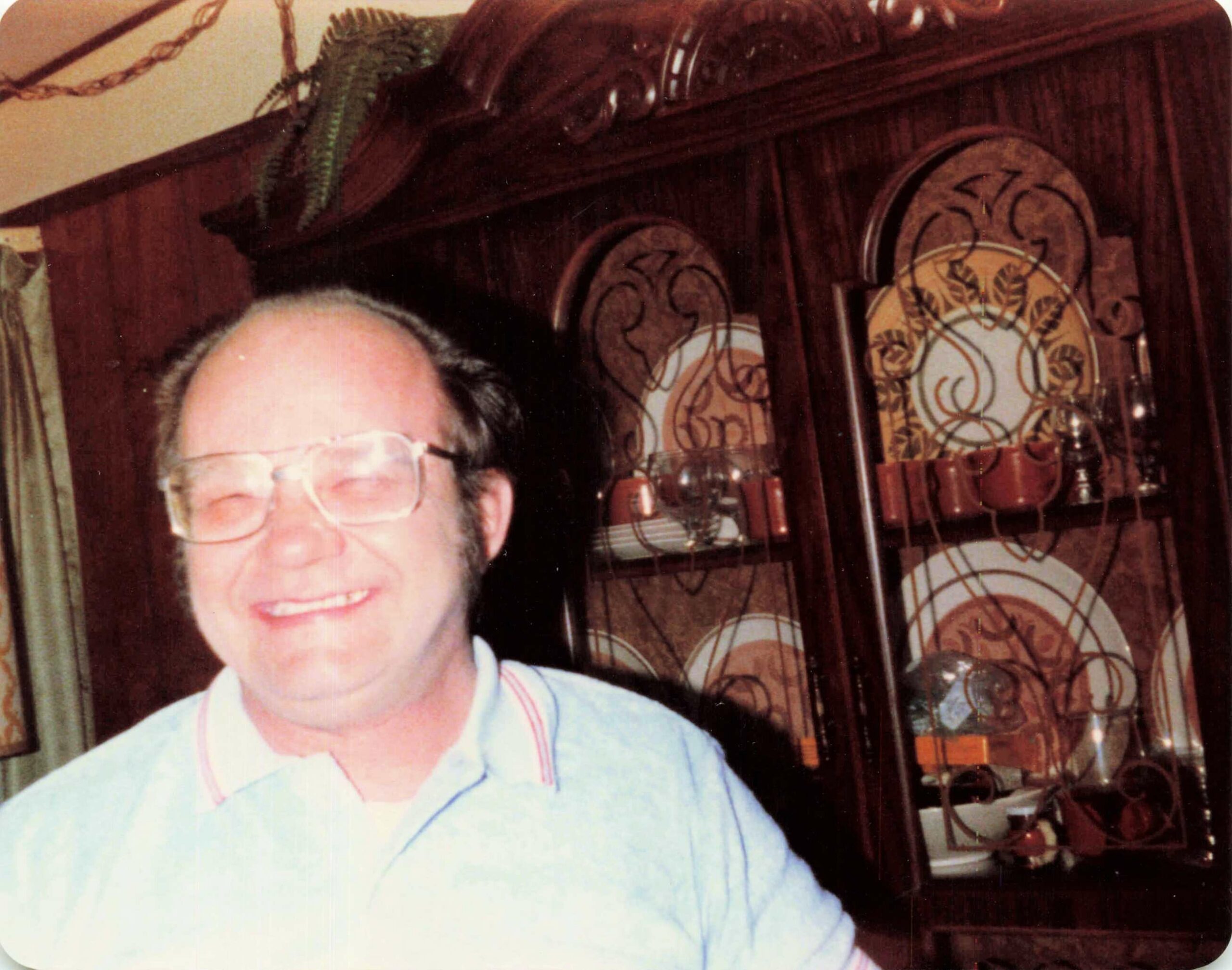 Partial portrait of a person wearing clear glasses, smiling, in front of a dark wooden cabinet with decorative plates and glassware behind him.