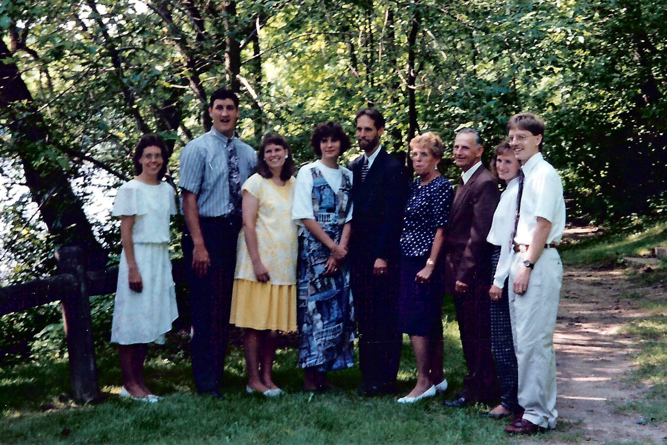 Group of eight adults posing together outdoors in a shaded park, smiling at the camera.