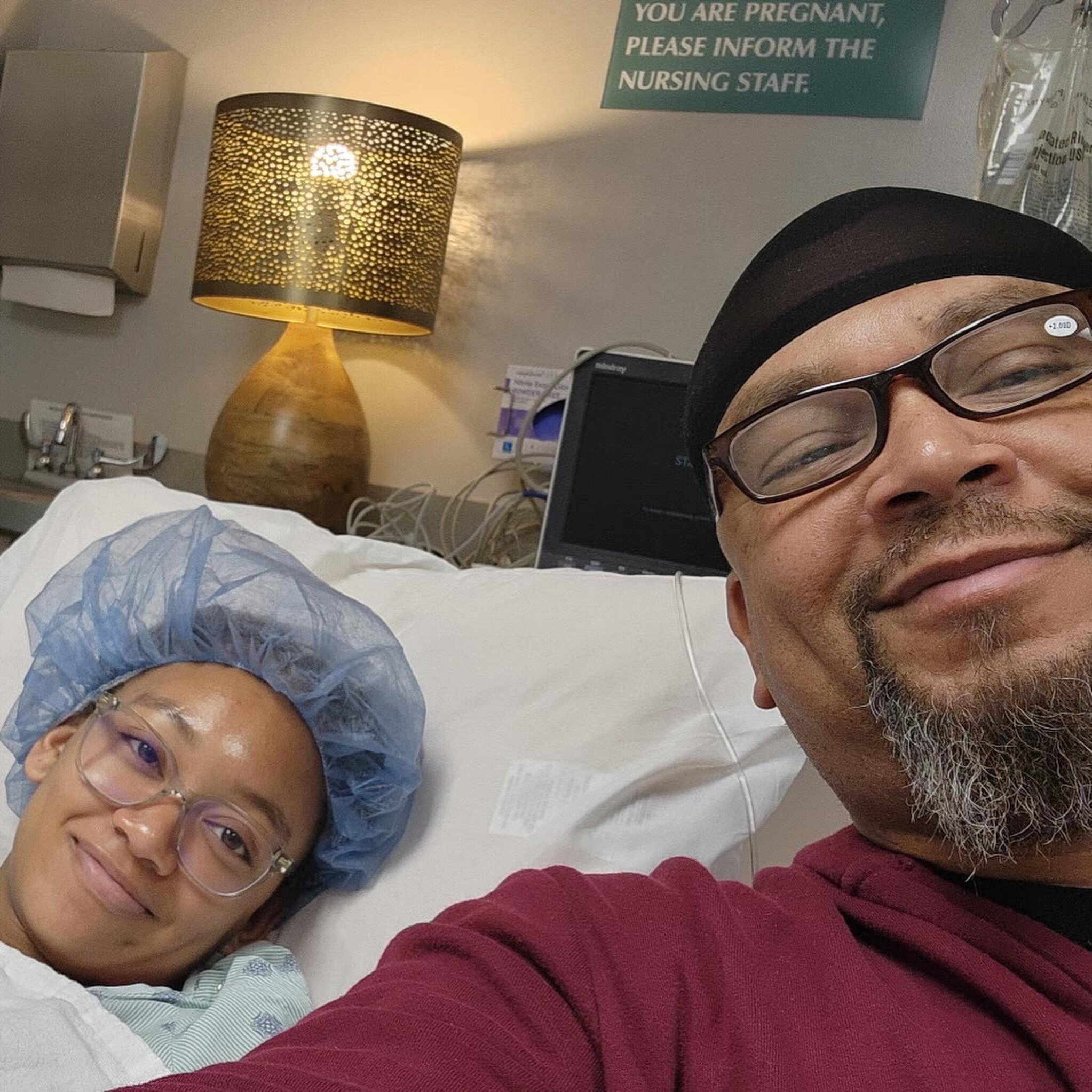 Selfie in a hospital room: man with glasses and stubble in a maroon sweater beside a patient wearing a blue surgical cap.