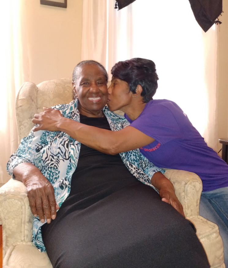 A woman in a teal and white patterned blouse sits in a chair while another person in a purple shirt kisses her cheek, both smiling and embracing.