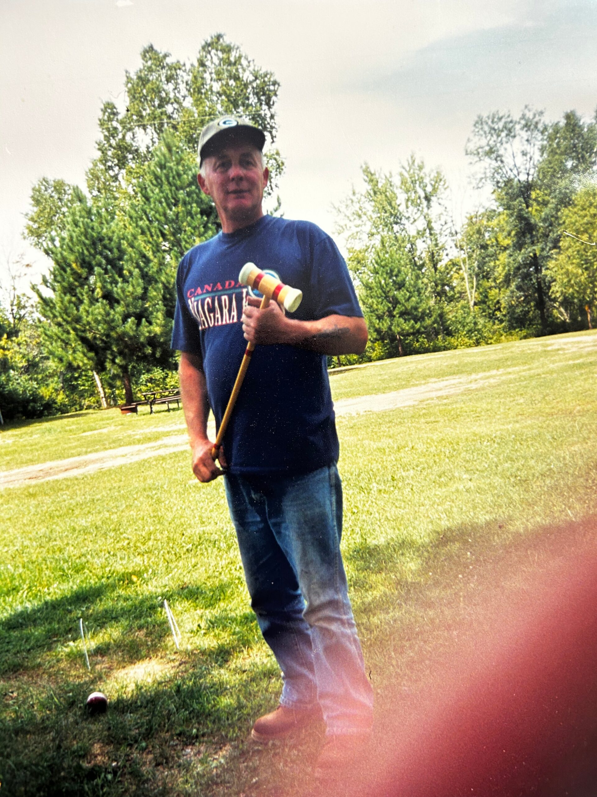 Man in a blue t-shirt and cap stands in a sunny park, holding a croquet mallet with a ball on the grass nearby.
