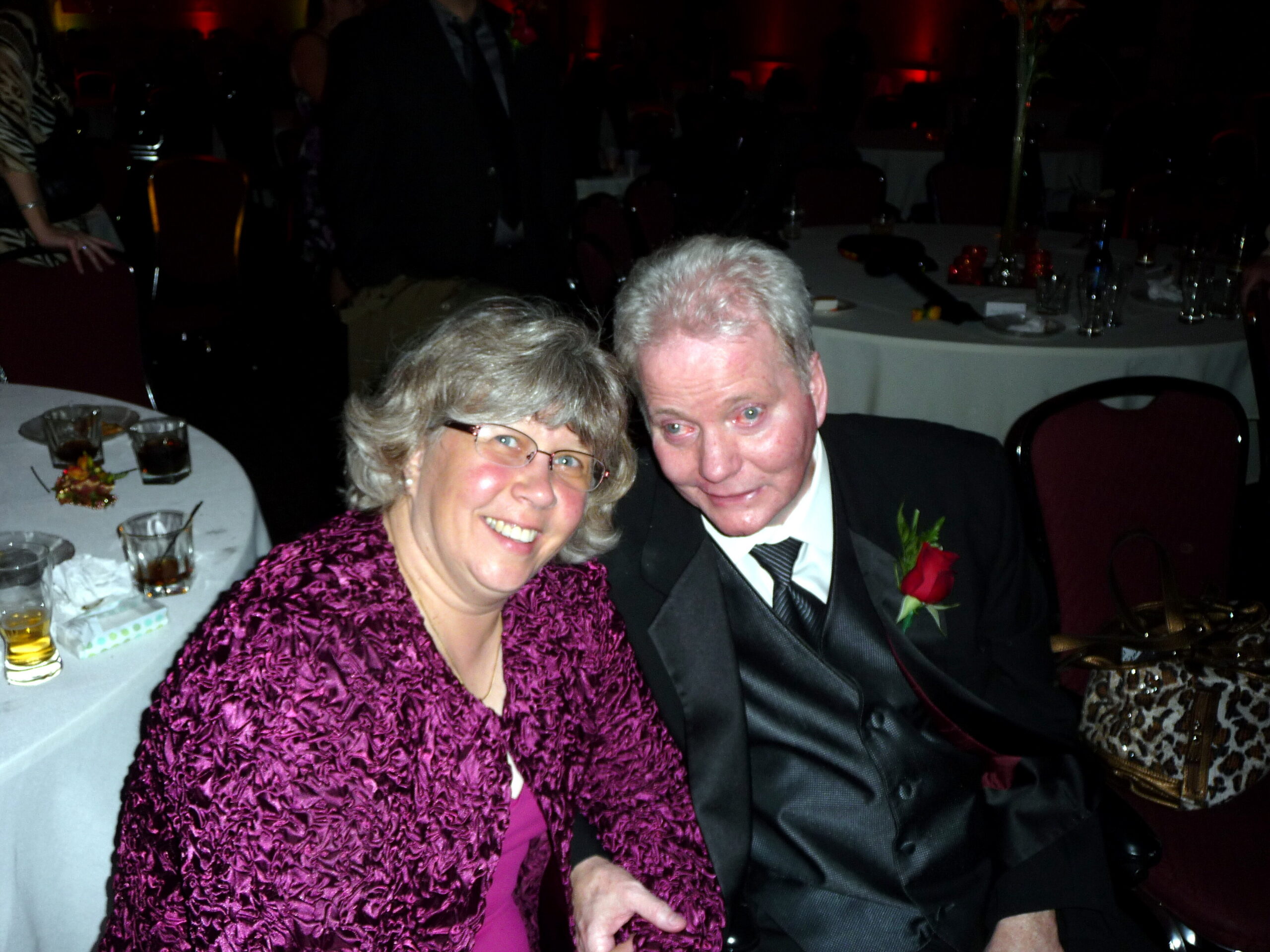 Smiling elderly couple at a formal gala, seated at a round table with drinks and a centerpiece in the background.