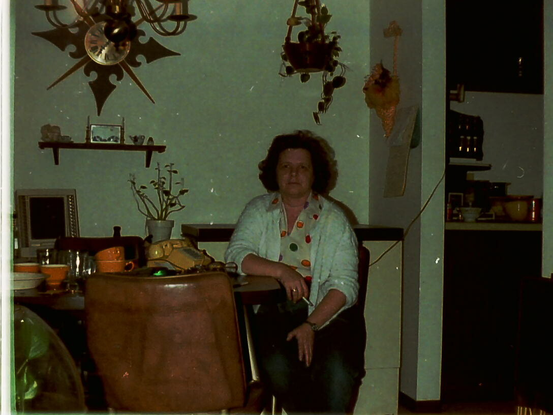Woman seated at a cluttered dining table in a retro-style home with a hanging chandelier overhead and green walls.