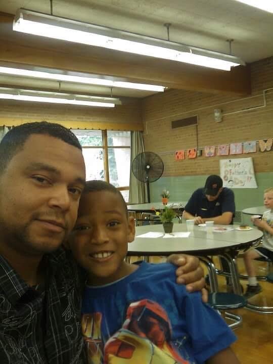 A man and young boy take a close selfie in a classroom or community center, smiling with others at tables in the background.
