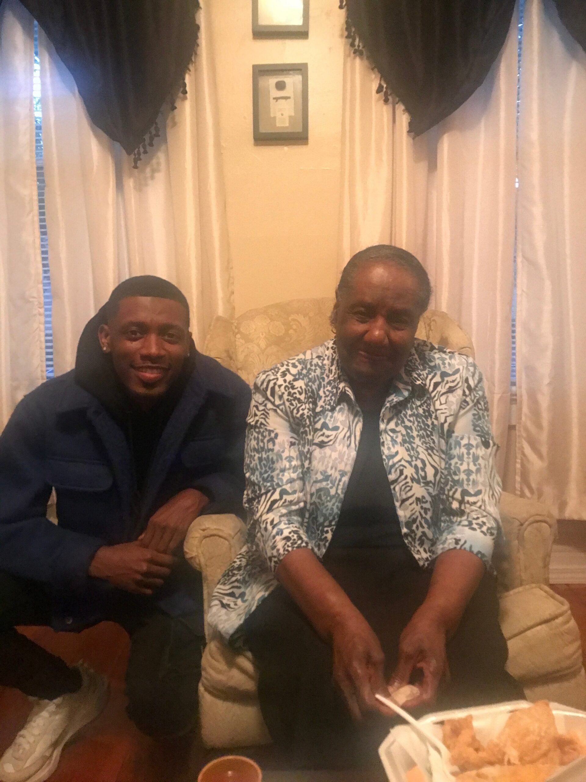 Two people sit on a beige sofa in a living room sharing a meal, with sandwiches on a tray in front.