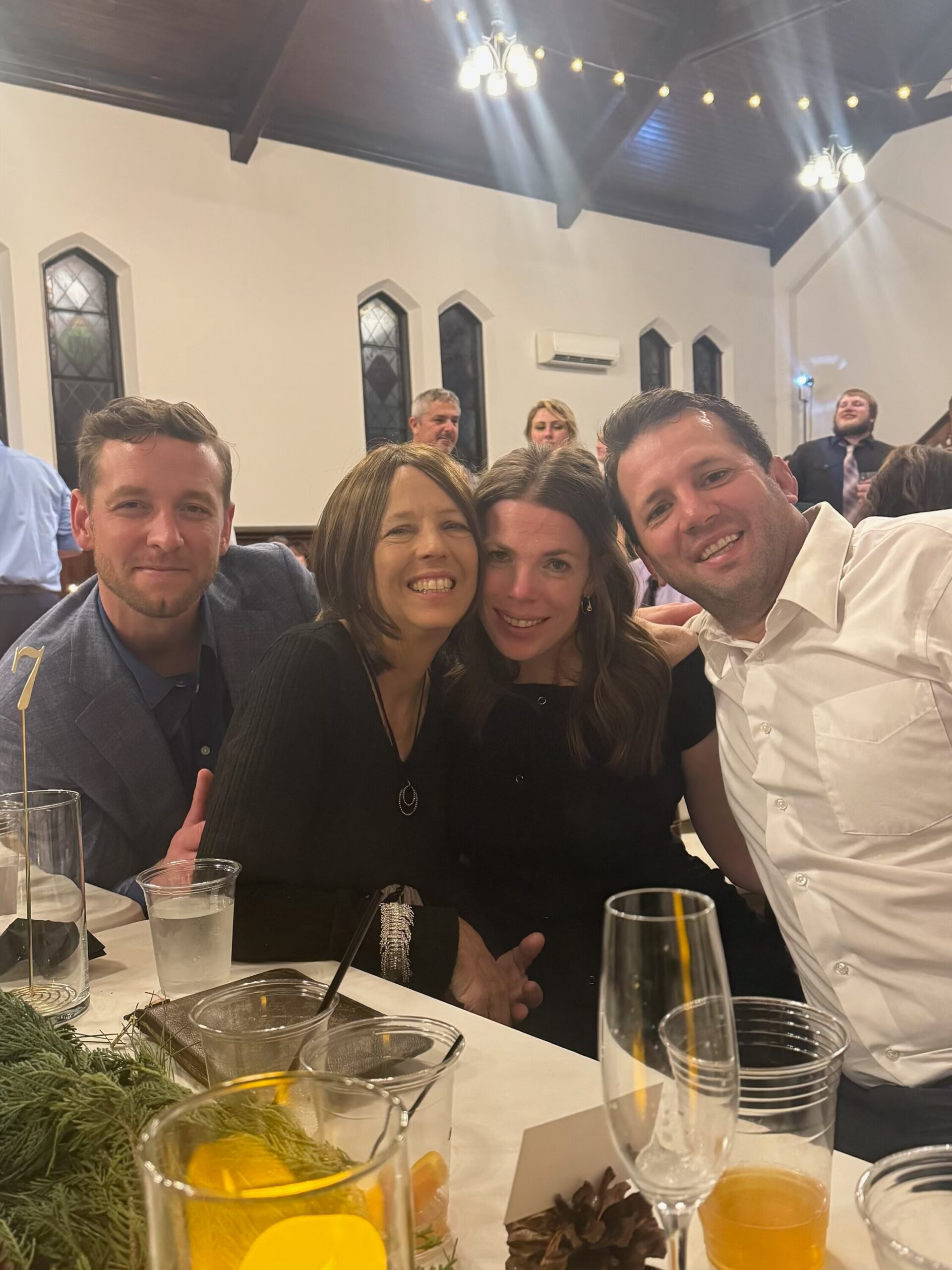 Four friends posing for a selfie at a dinner table during a celebration, with drinks and greenery in the foreground inside a decorated venue.