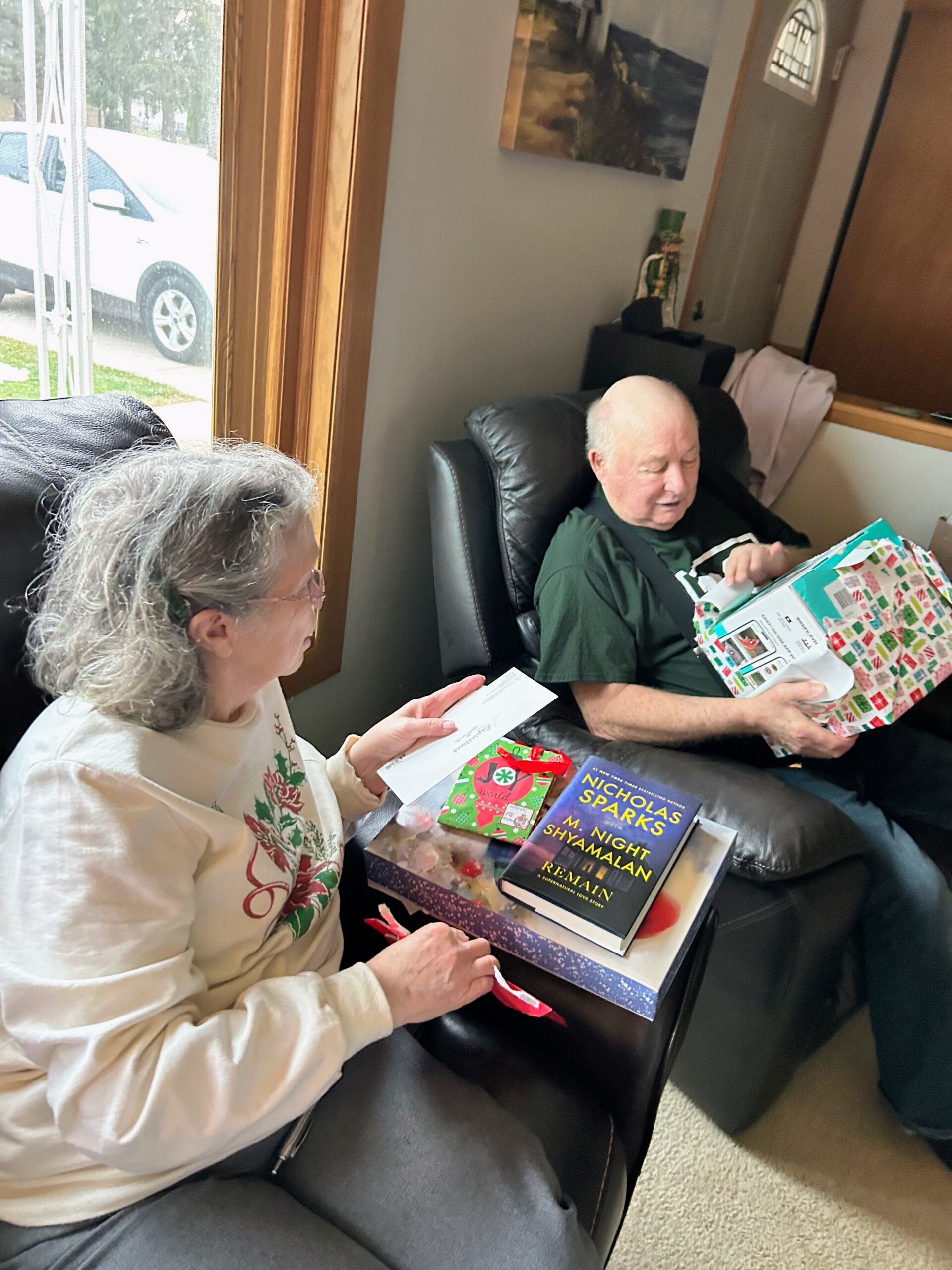 Two seniors sit in a living room, opening wrapped gifts; a man in a green shirt looks at a present while a woman nearby holds a card.