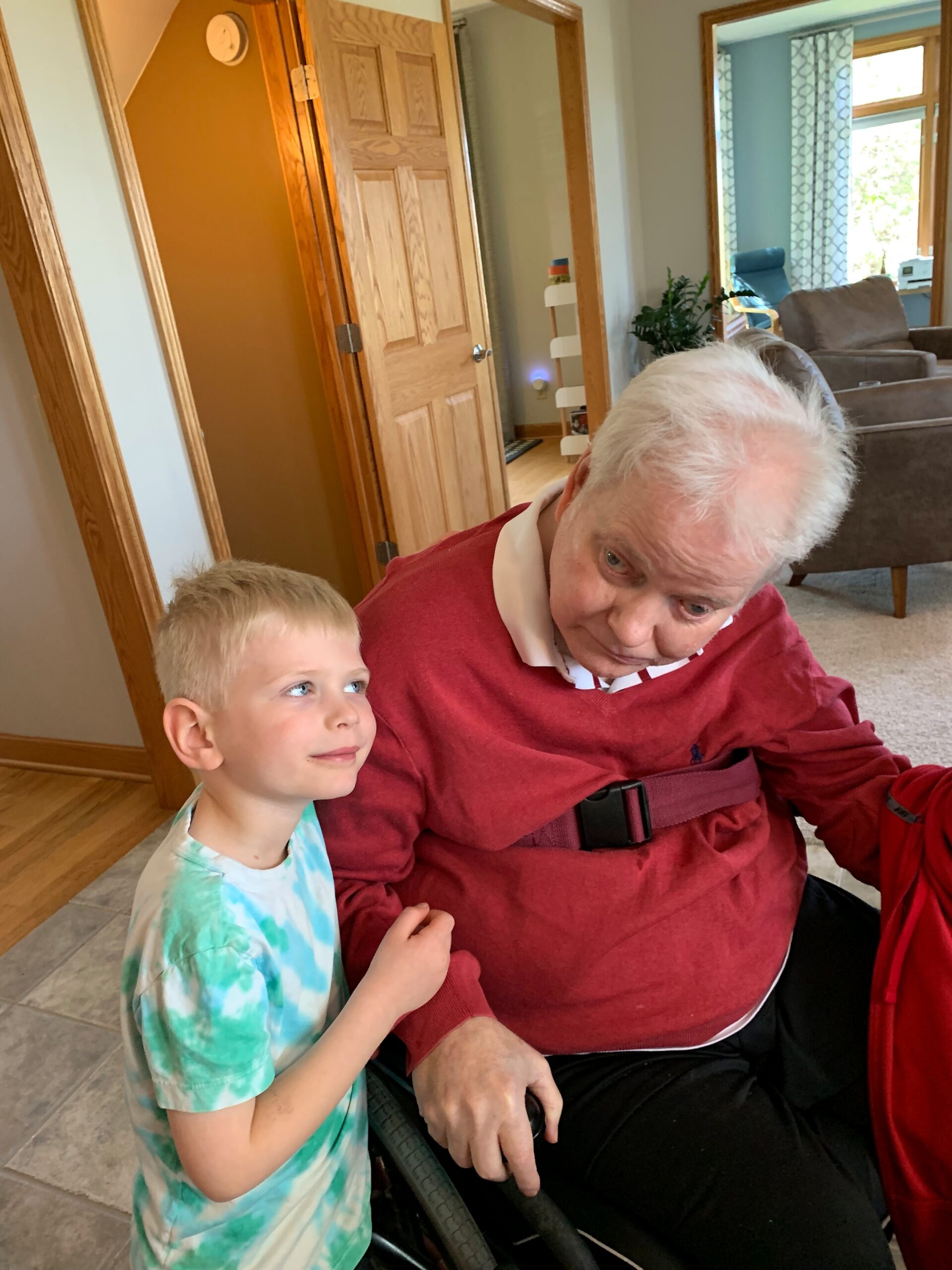 Young boy in a tie-dye shirt stands beside an elderly man in a red sweater, seated in a wheelchair inside a home.