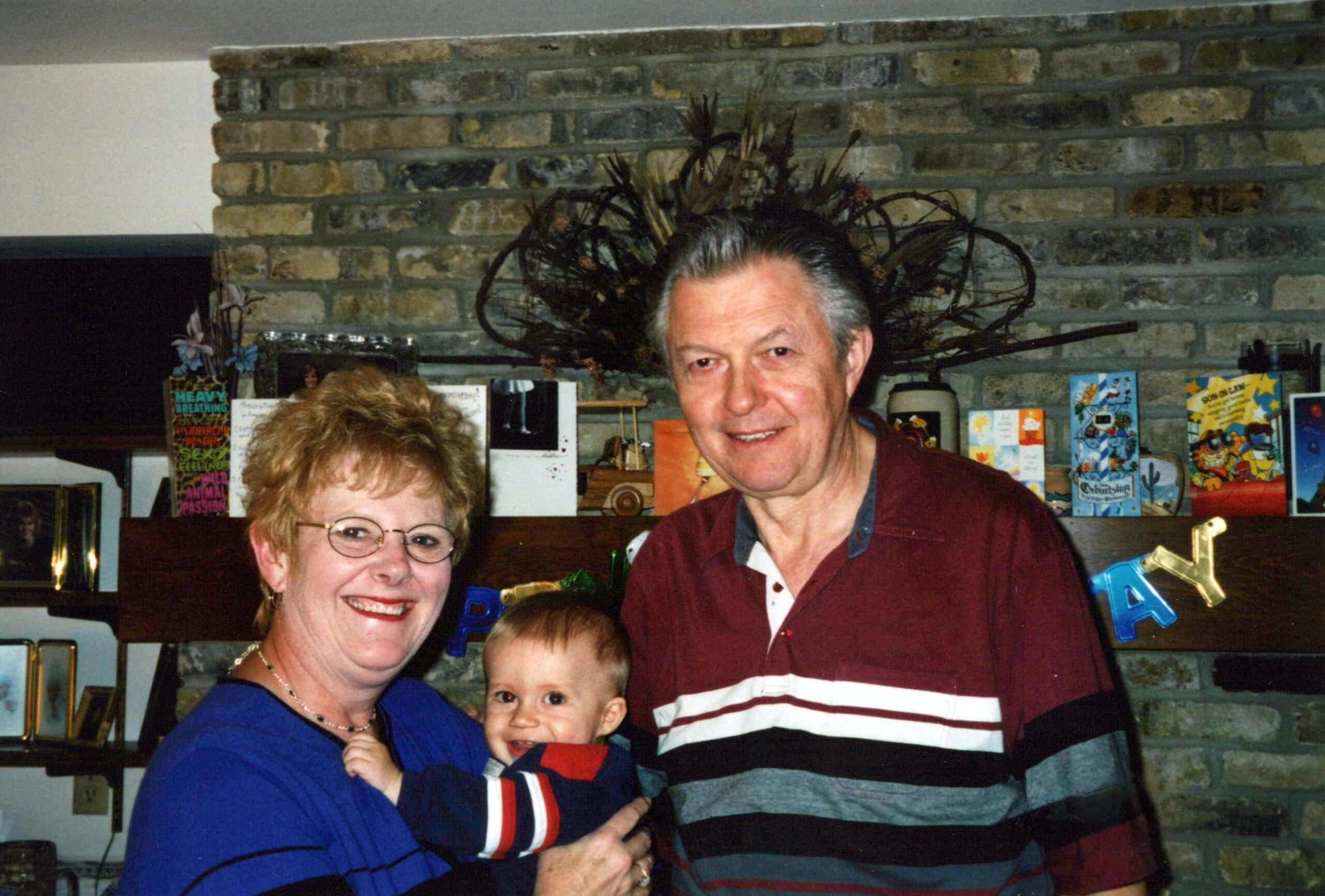 Smiling older woman, man, and baby posing together indoors in front of a brick wall with cards and decorations in the background.