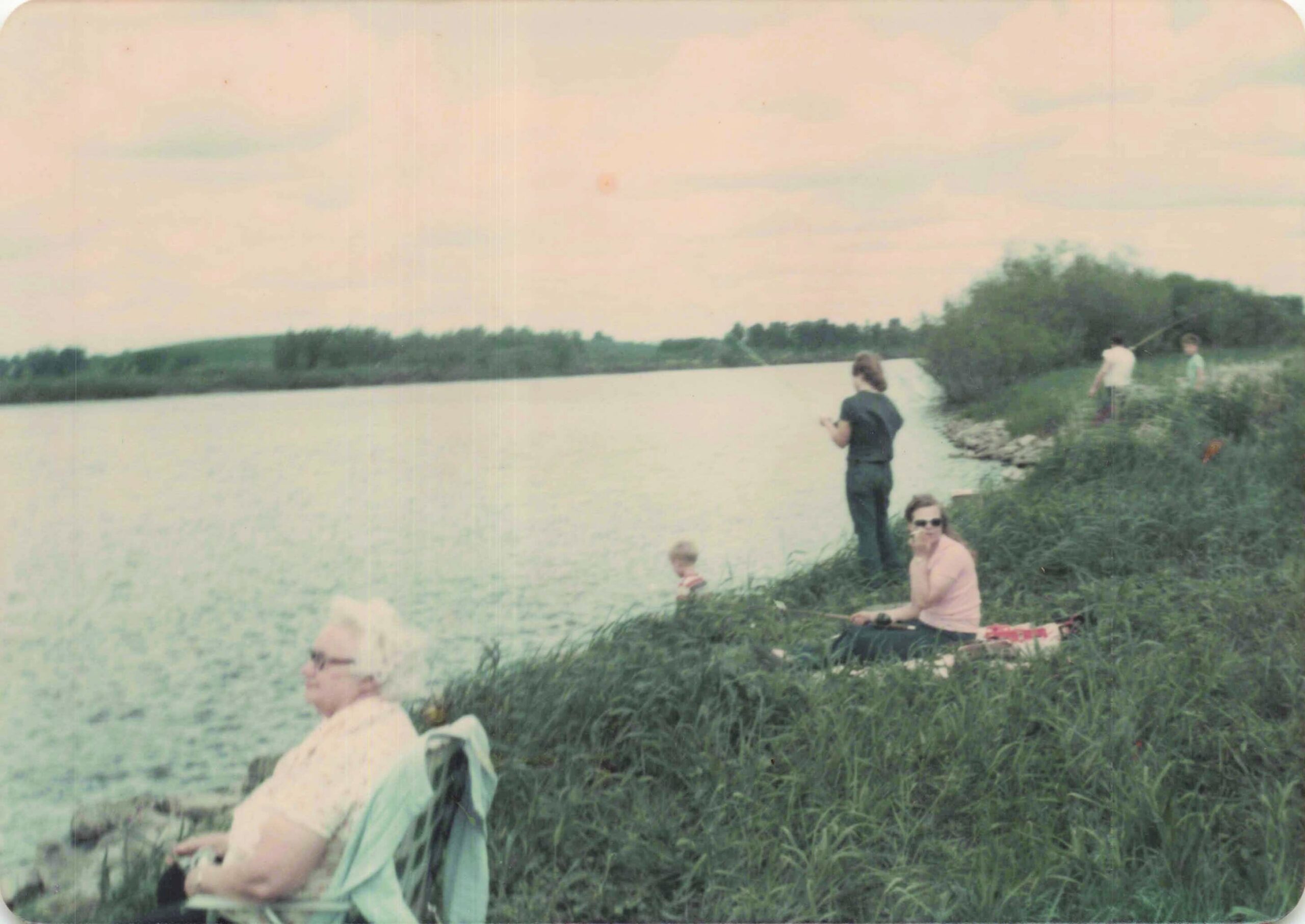 People fishing along a grassy riverbank by a calm lake, with a child in the foreground.