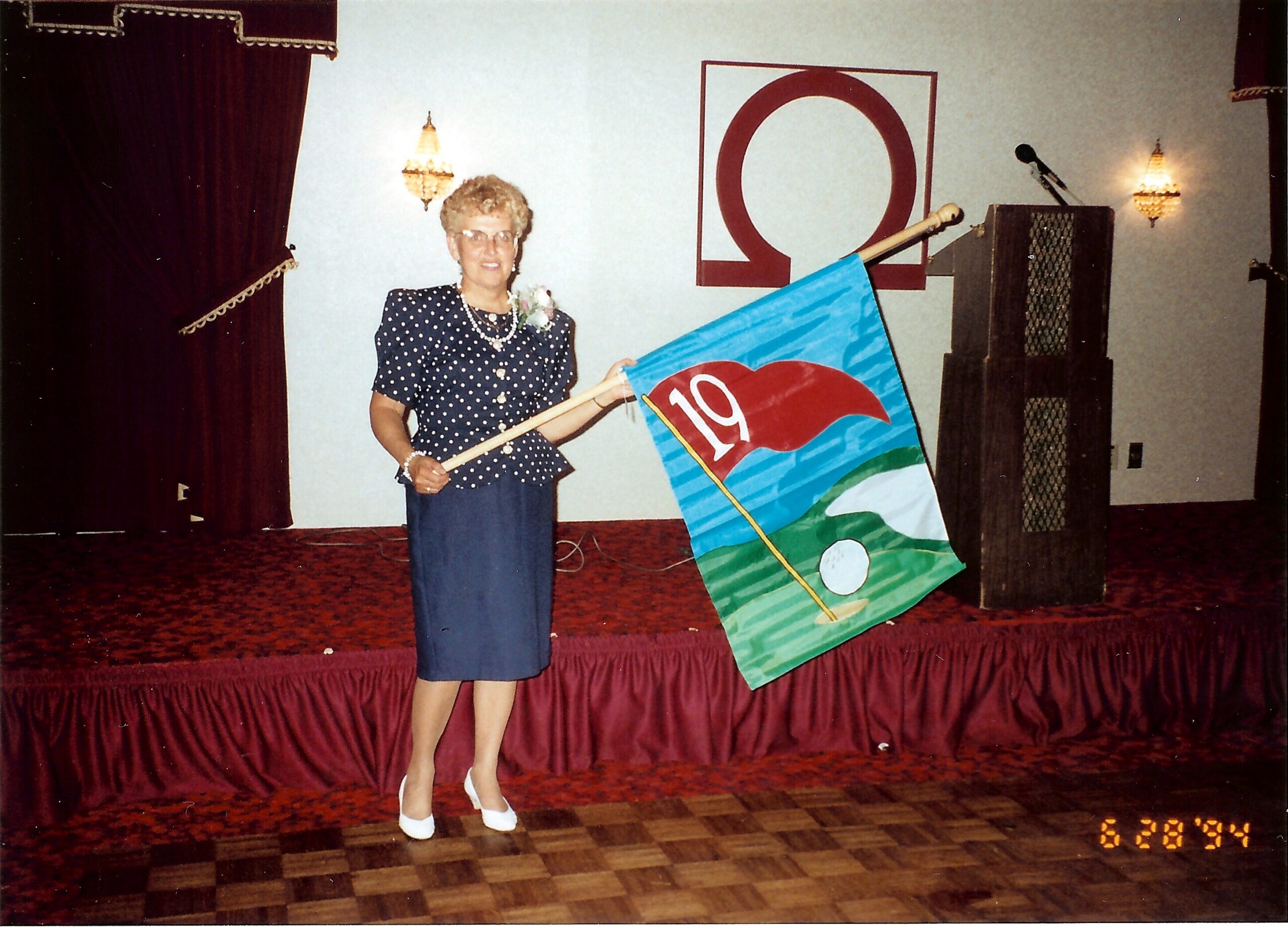 Older woman in a navy polka-dot blouse with pearls on a stage, holding a blue flag with a bold red circle emblem.