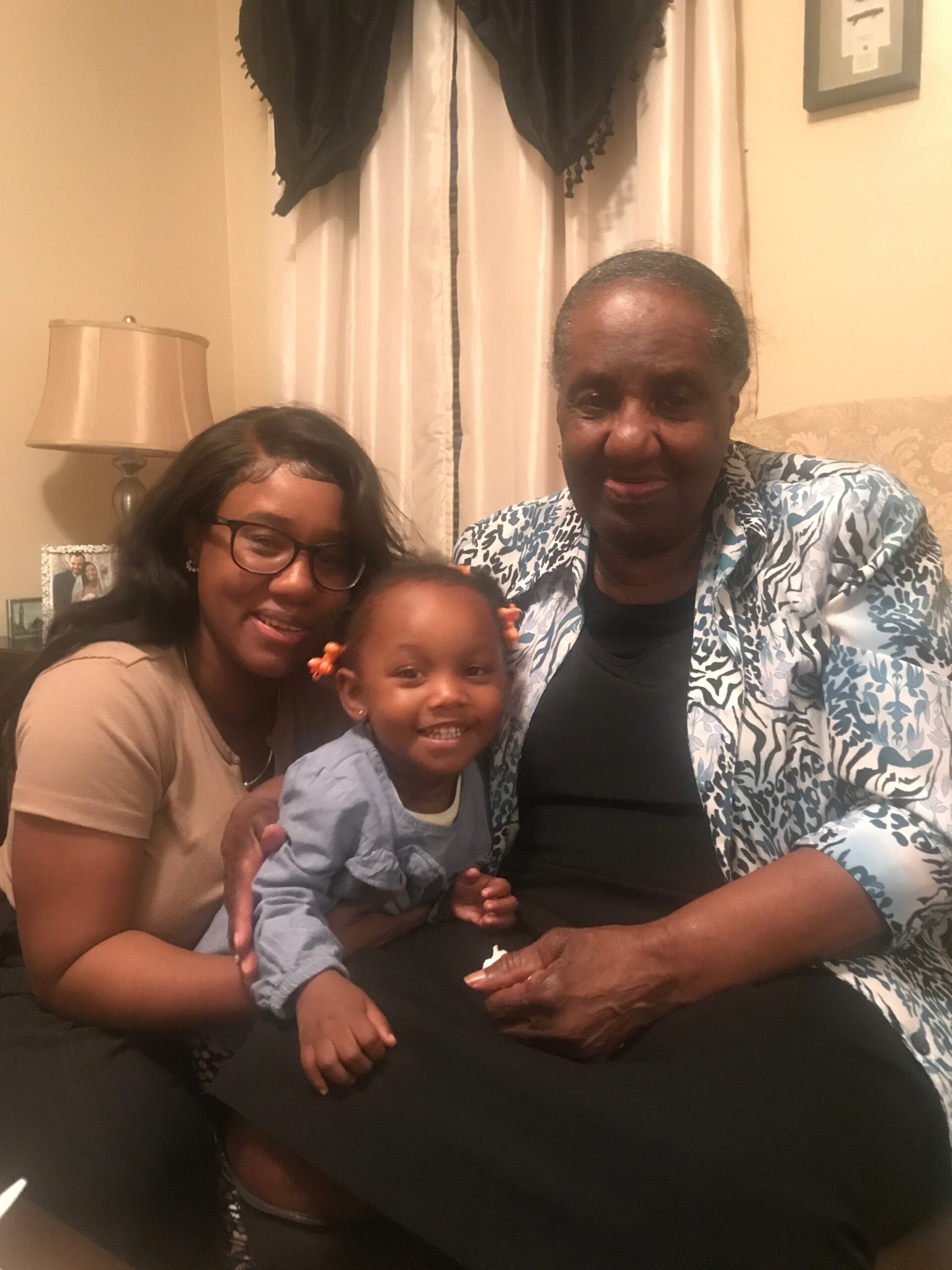 A trio of family members sits on a couch for a warm living-room portrait: a grandmother on the right, a woman with glasses on the left, and a smiling child in the middle holding hands with both adults, with a lamp and curtains in the background.