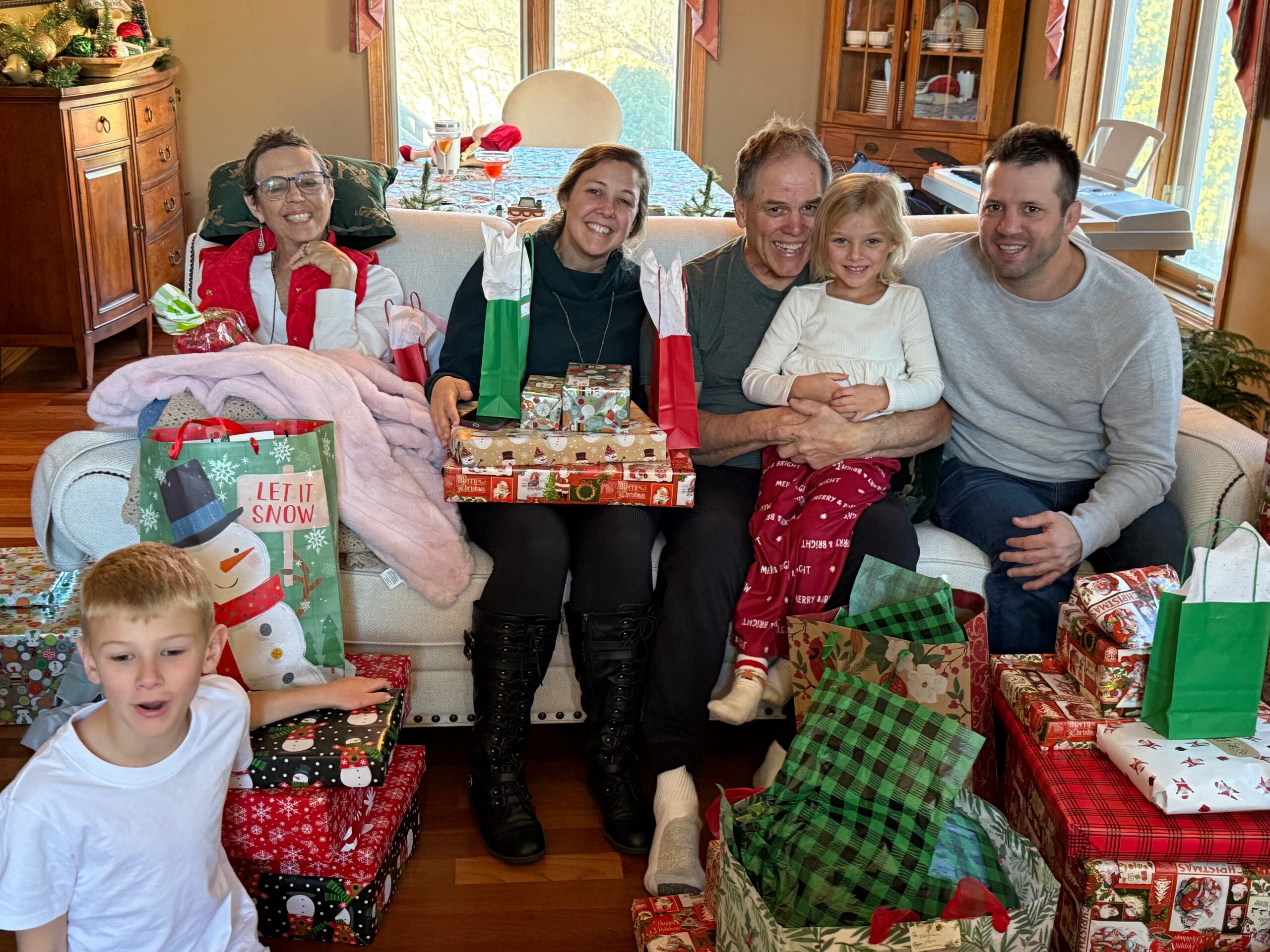 A multi‑generational family sits on a couch surrounded by wrapped Christmas gifts, smiling during a holiday gathering.