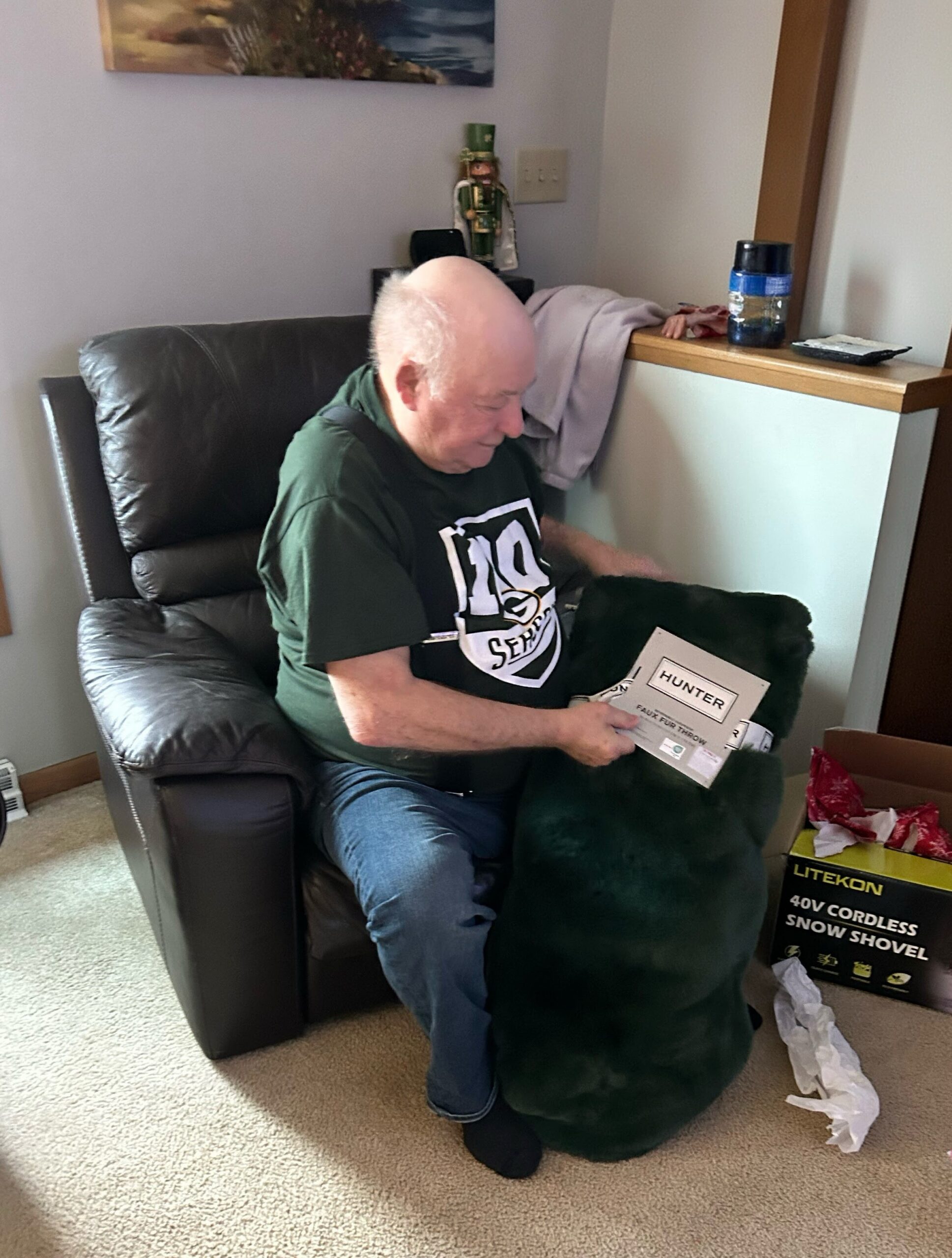 An older man sits in a leather chair, wearing a green shirt, looking down at a magazine on his lap with a large dark furry dog resting on his legs.