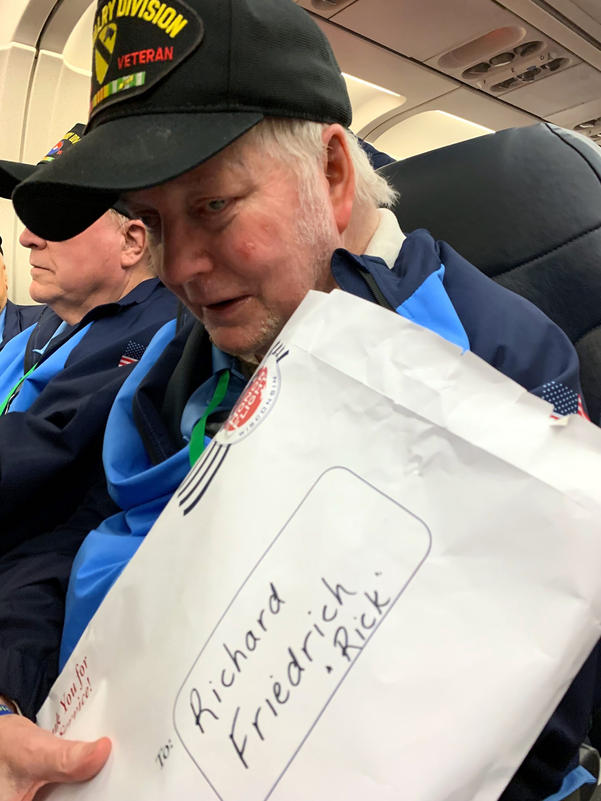 Older man wearing a black cap labeled 'Veteran' and a blue jacket sits on an airplane, holding a large white paper bag with handwritten text visible on the front.