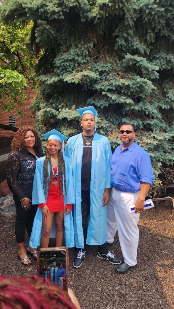 Group of four posing outdoors for a graduation photo: two graduates in blue gowns and caps, flanked by a woman and a man in smart casual clothing, plus another man holding a rolled diploma.