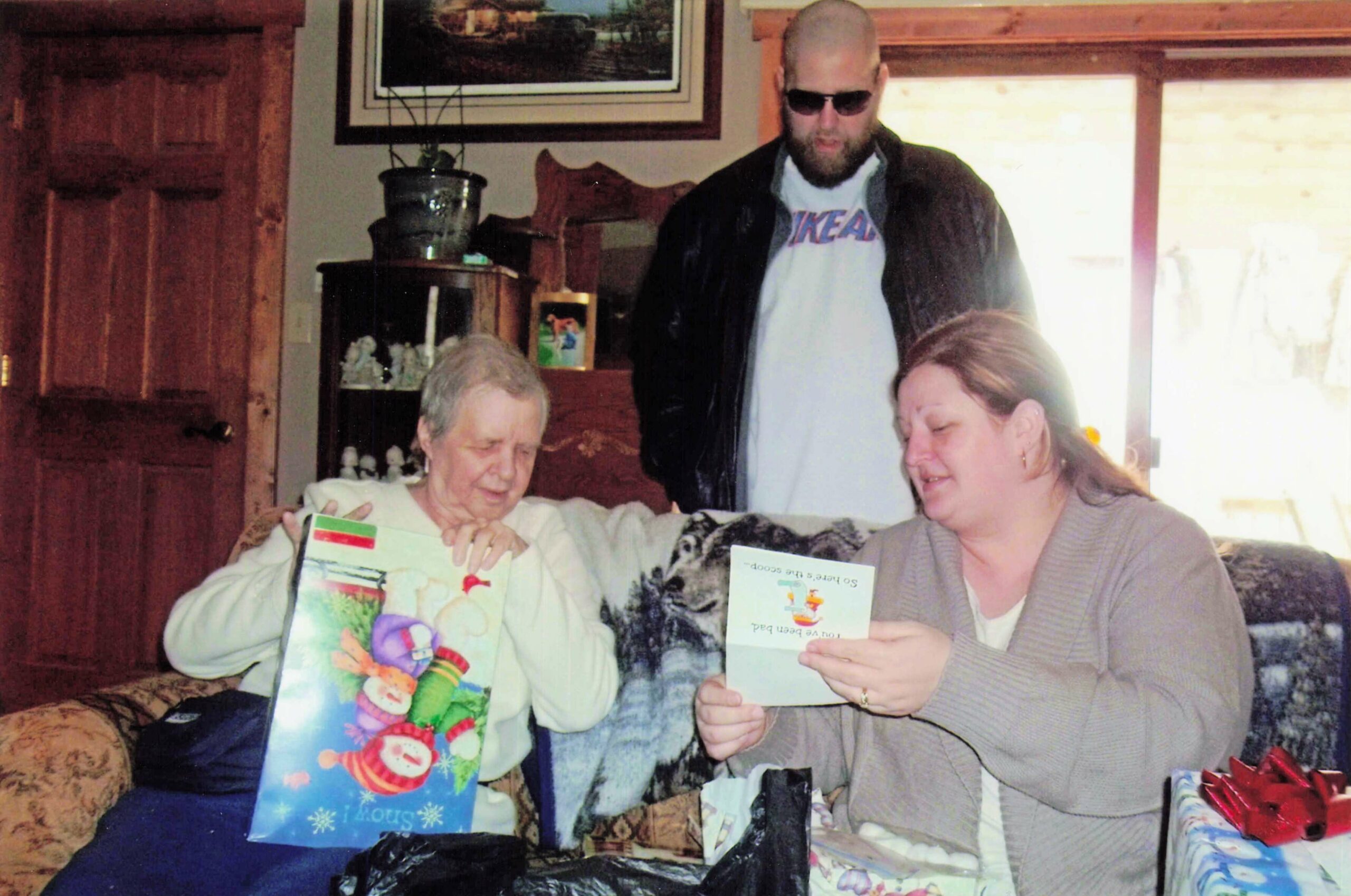 Two women open gifts on a sofa while a bearded man in sunglasses stands behind them in a living room.