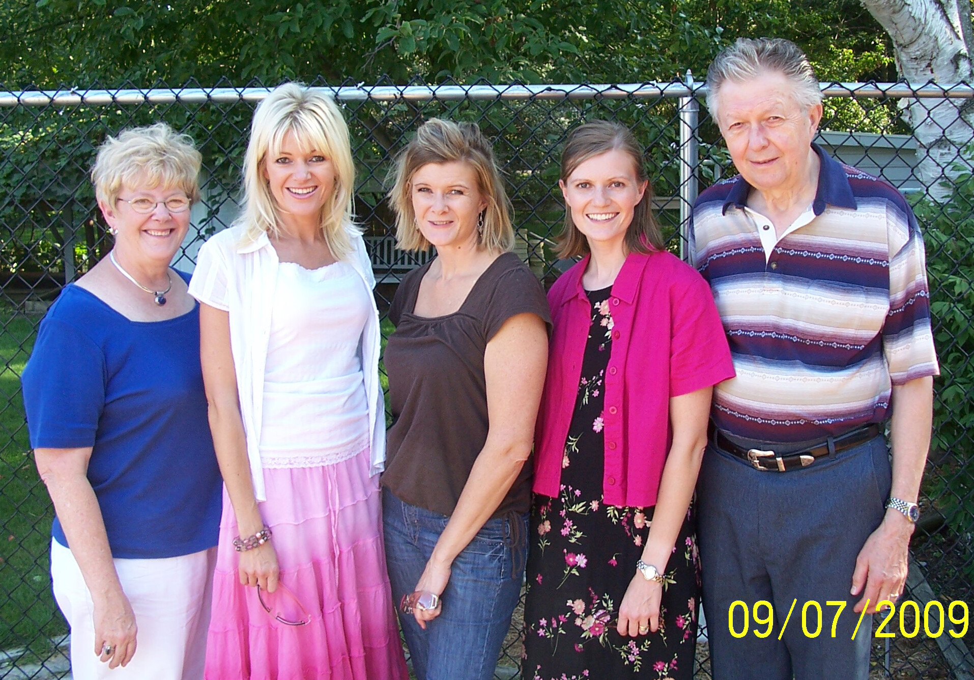 Five adults posing together outdoors in front of a chain-link fence, smiling at the camera, with a yellow date stamp visible in the bottom corner.