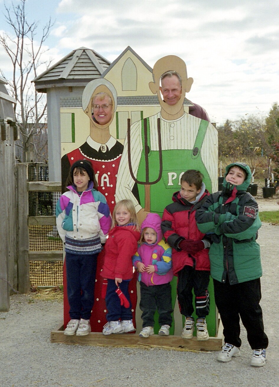 Children pose behind life-size cutouts of a farmer and his wife on a farm, all smiling at the camera behind the face holes on the cardboard figures