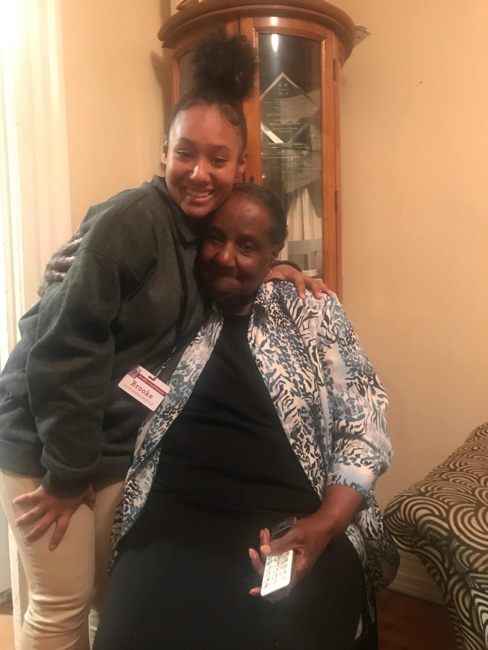 Young woman in a dark hoodie hugs an older woman in a blue-and-white patterned shawl, seated in a living room with a wooden cabinet behind them.