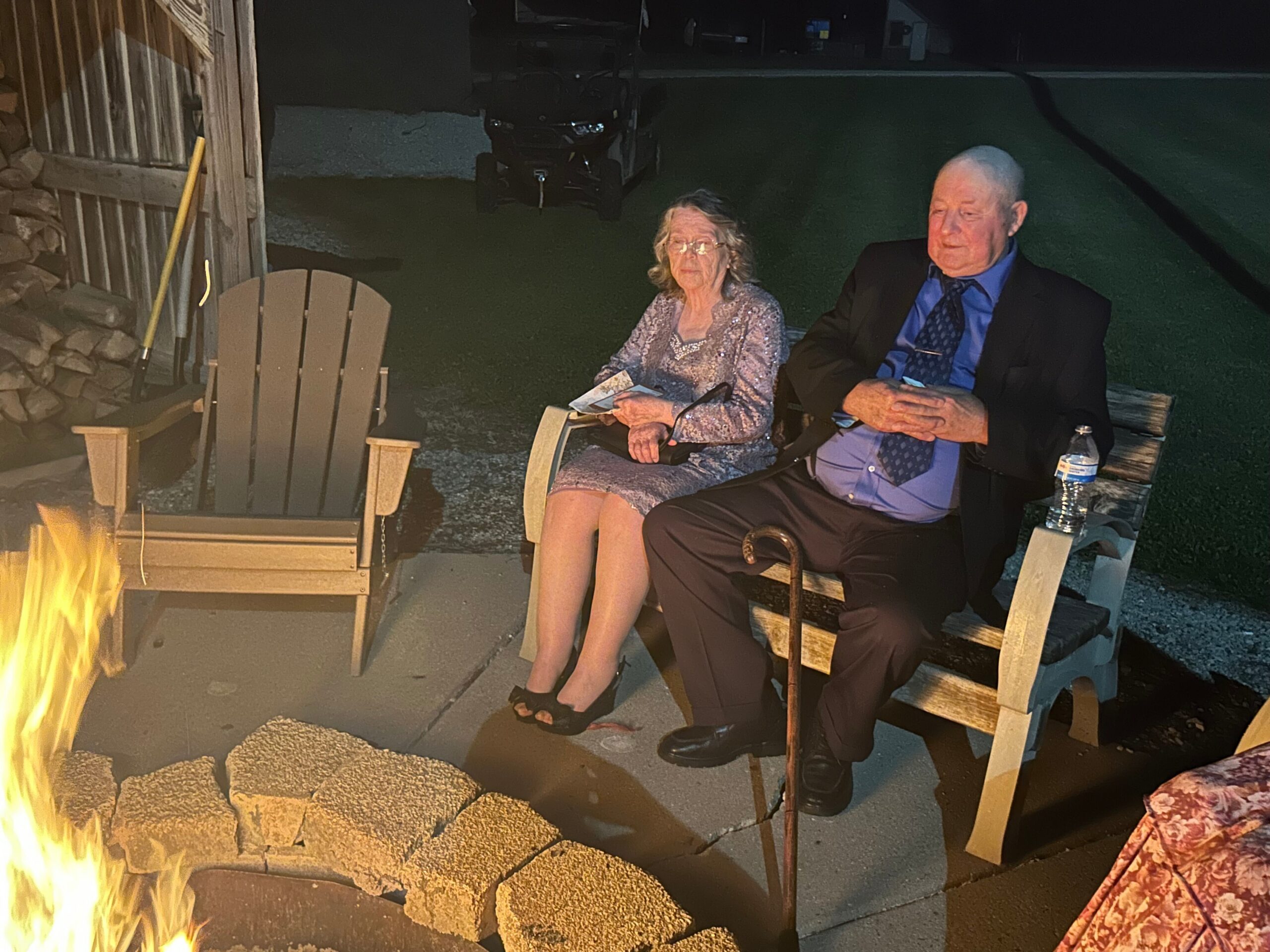 Elderly couple seated in wooden chairs by a lit backyard fire pit at night, flames in the foreground and a bottle on the table nearby.