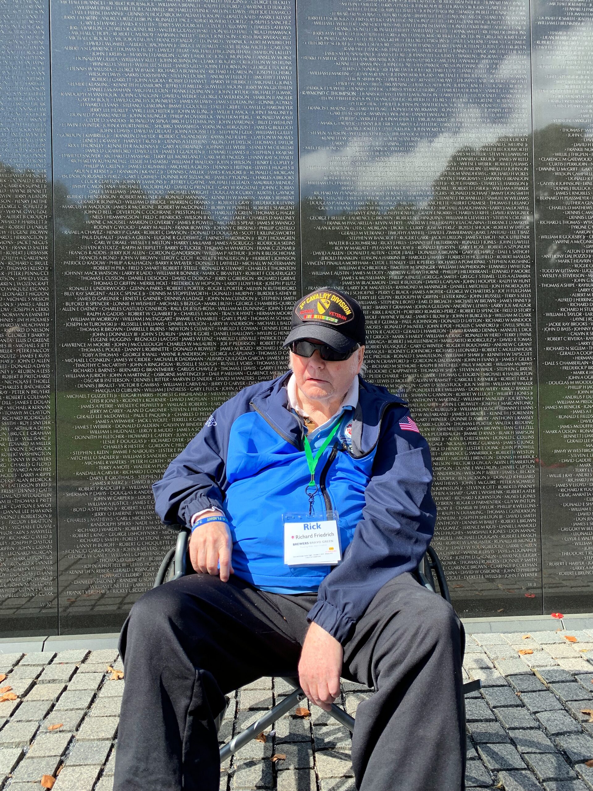 Person in a wheelchair in front of a wall covered with names engraved in metal; wearing a navy jacket, blue shirt, sunglasses, cap with Cavalry Division patch, and a green lanyard badge