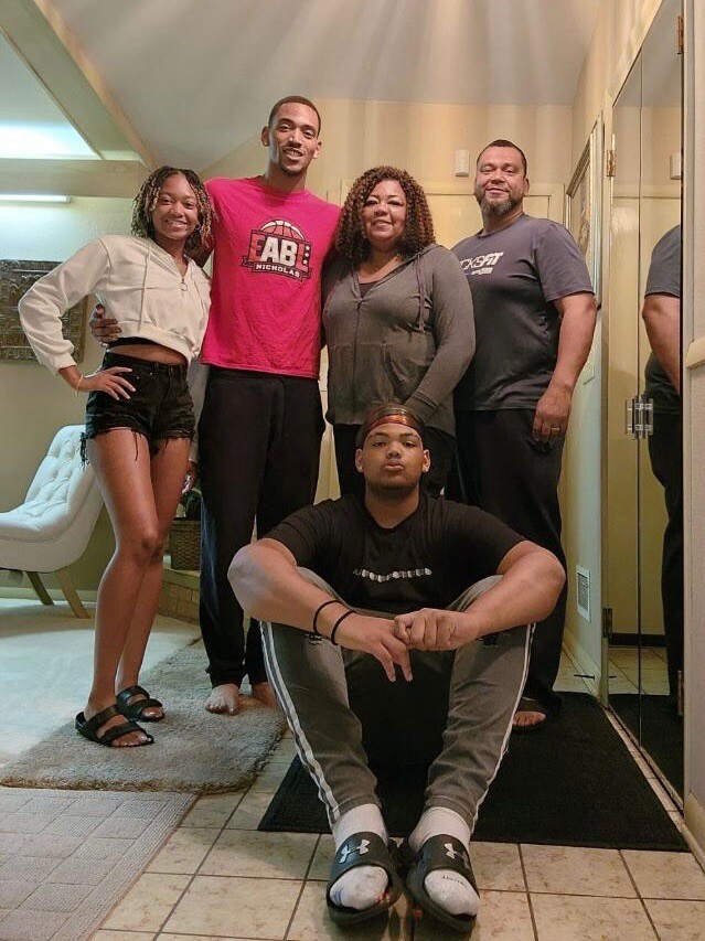 Group of five people posing together in a hallway, one seated on the floor in front wearing black and gray athletic wear.