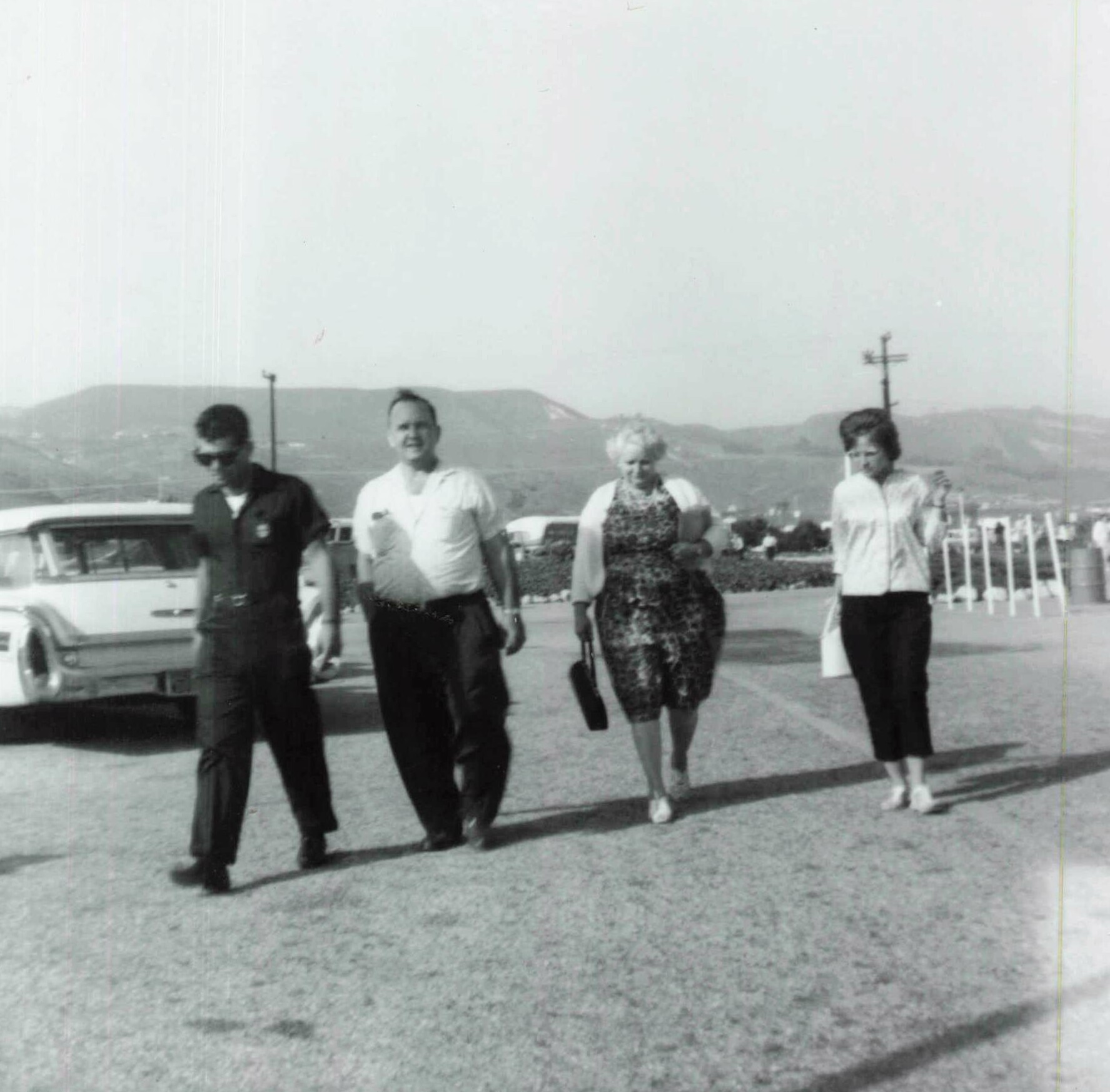 Four adults walking toward the camera in a parking lot with mountains in the background; casual clothing, vintage photo style.