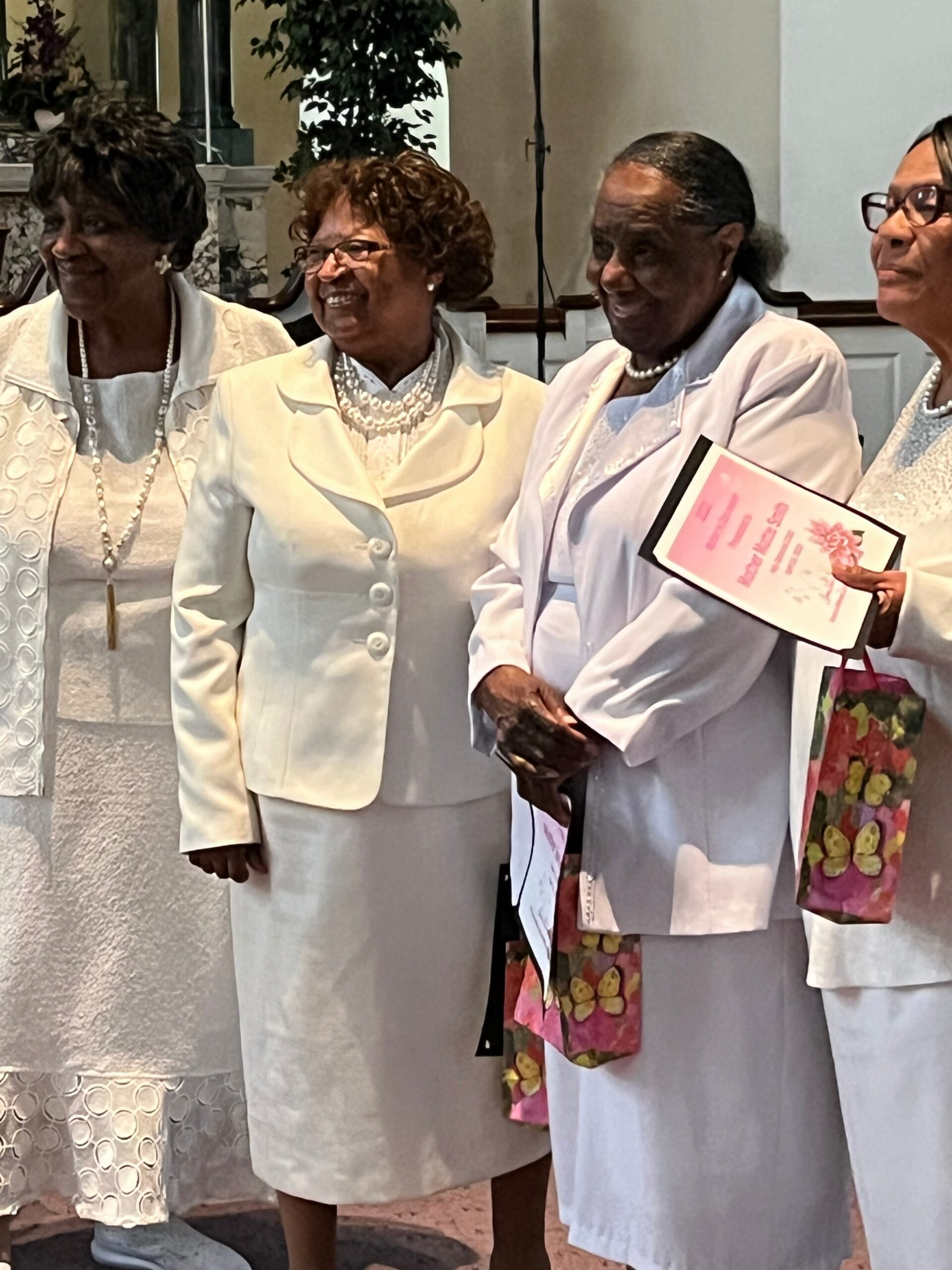 Group of four elderly women in pale cream outfits smiling together at an event, holding a pink floral gift bag and a program booklet.