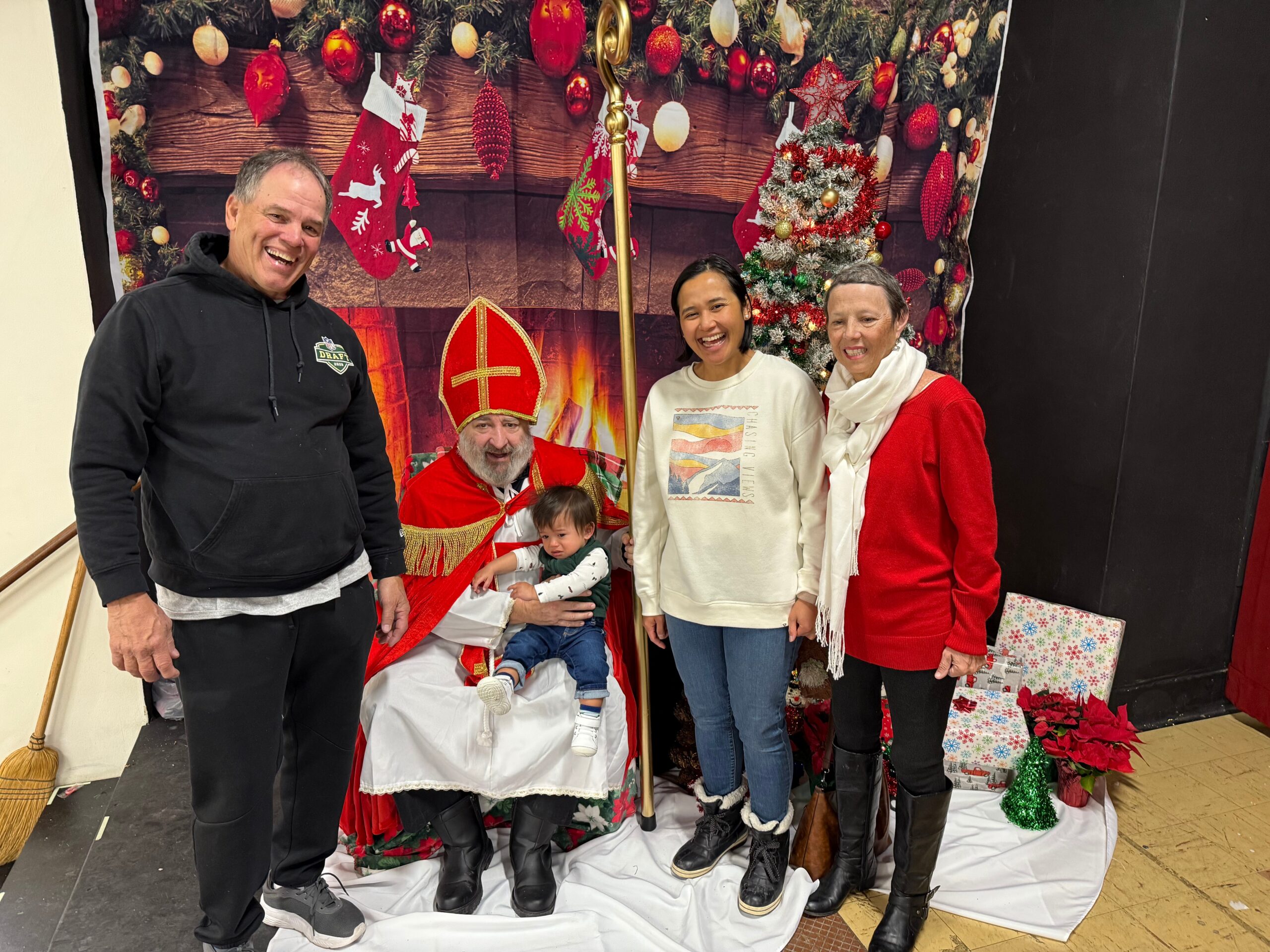 Group of adults and two children posing with Santa in a festive Christmas backdrop with stockings, tree, and wrapped gifts nearby.