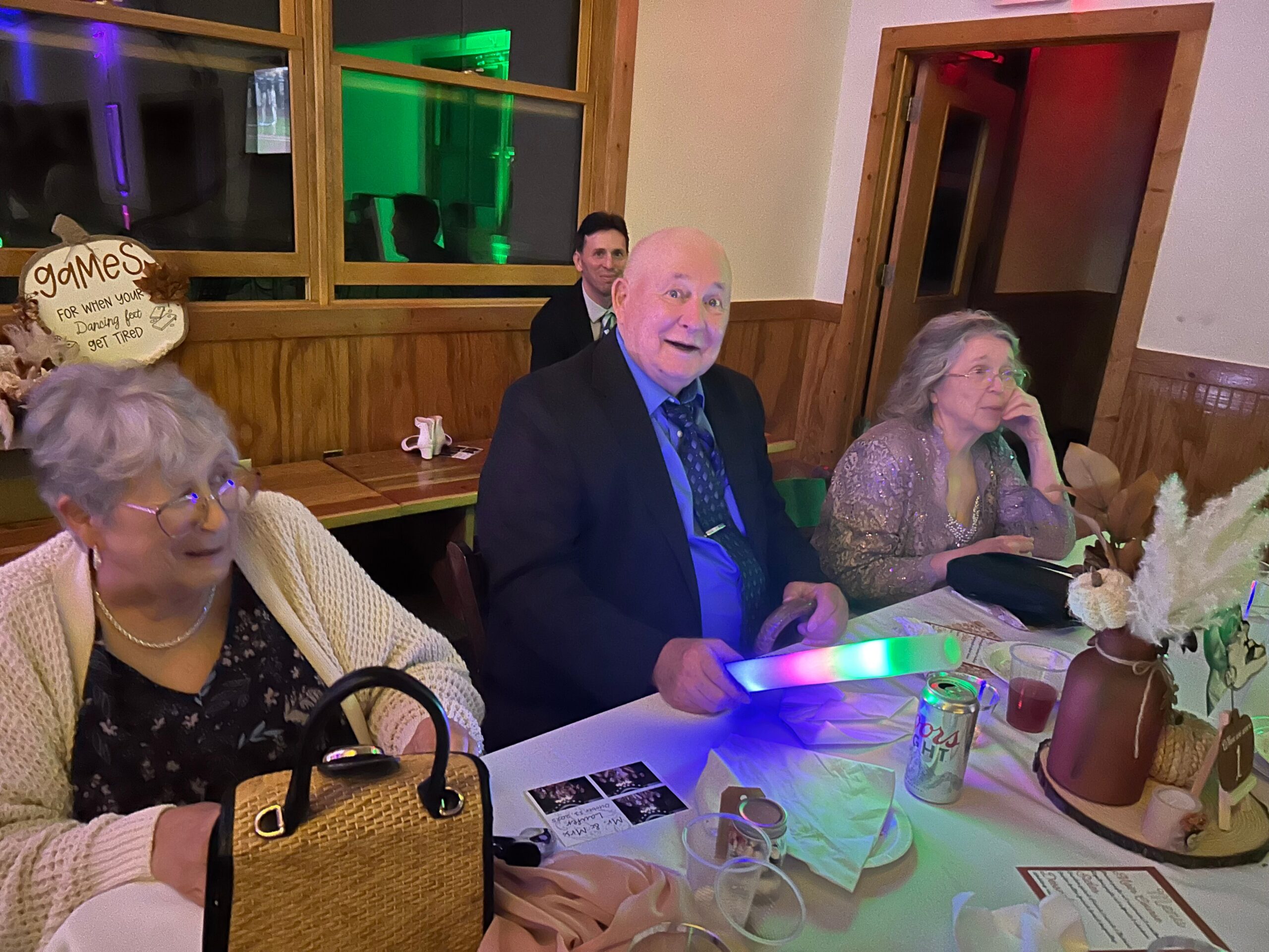 Group of seniors seated around a table at a party, smiling and chatting under colorful lights with drinks and games on the table.